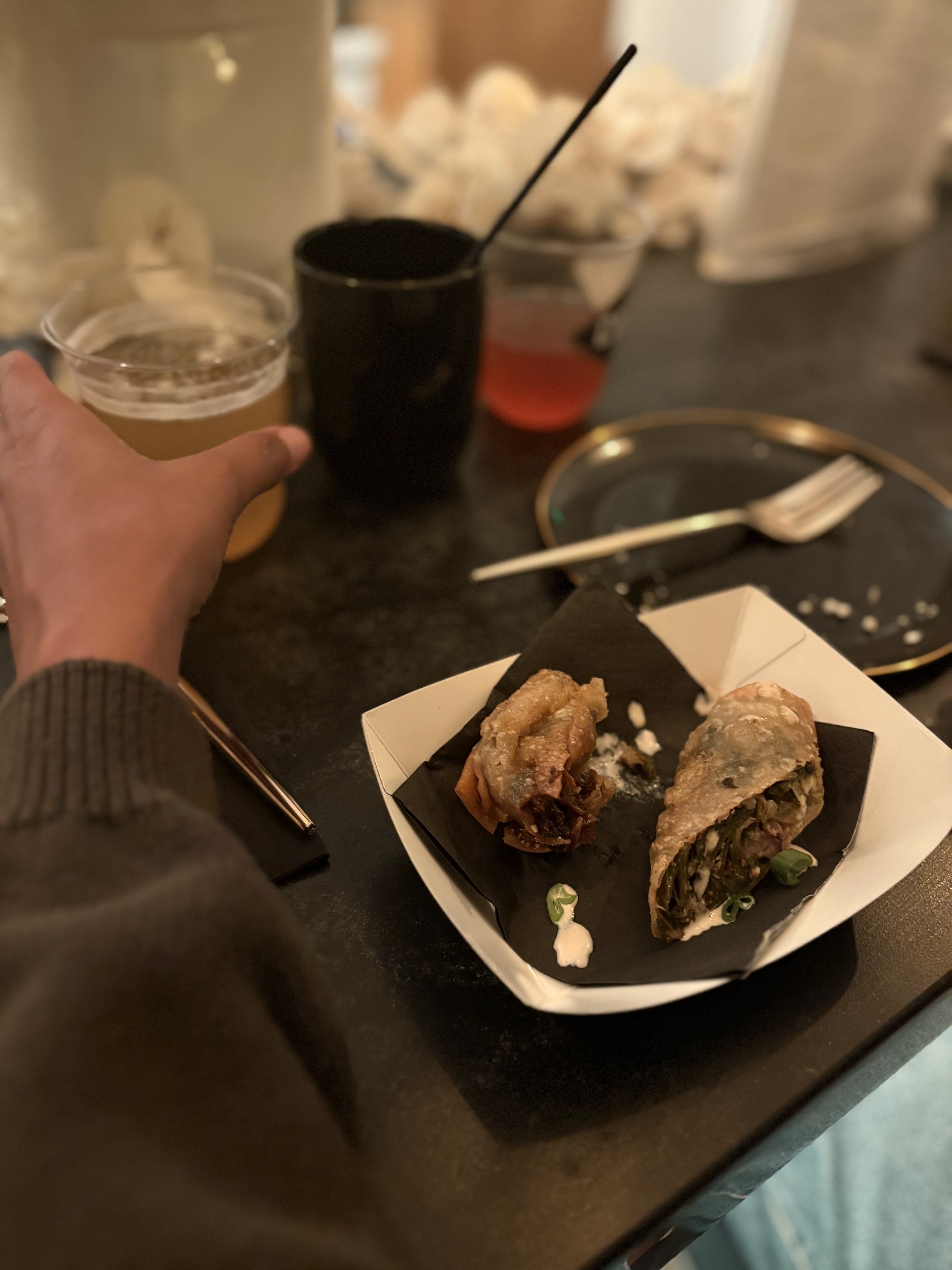 Close-up of a paper food tray containing two egg rolls, with a green onion garnish, set on a black table. Blurred background shows beverage cups, a fork, and a small bowl with a pink-colored drink.