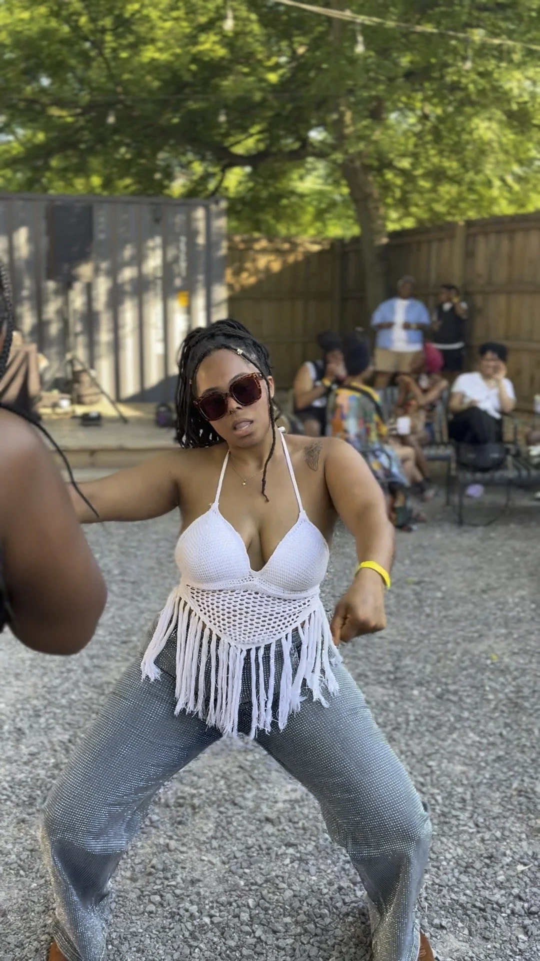 A woman with dreadlocks wearing sunglasses, a white crochet top with fringe, and sparkly pants dancing outdoors at a gathering.