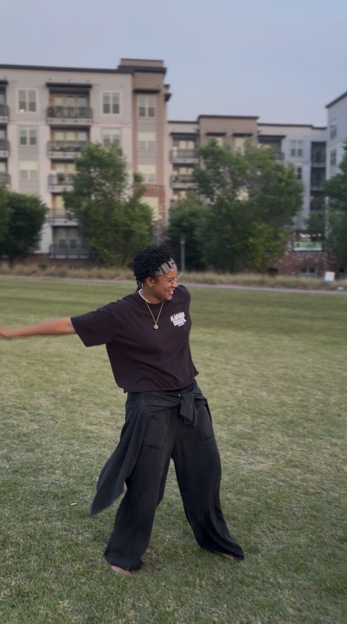A woman standing on grass in an outdoor park, smiling, with her arms stretched out to the sides, wearing a black T-shirt, black wide-leg pants, a headband, and necklace. There are apartment buildings and trees in the background.