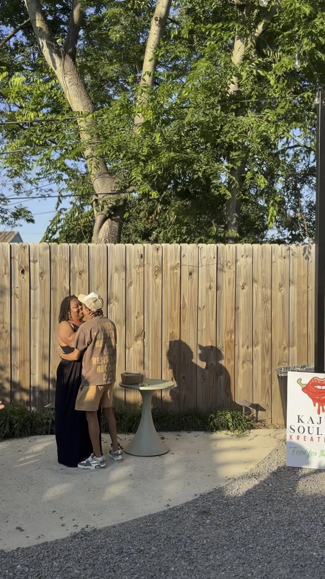 Two people hugging and sharing a kiss outdoors at a venue, with a wooden fence and pubic logo sign nearby. Shadows of the two individuals are cast on the fence, with large trees and a clear blue sky in the background.