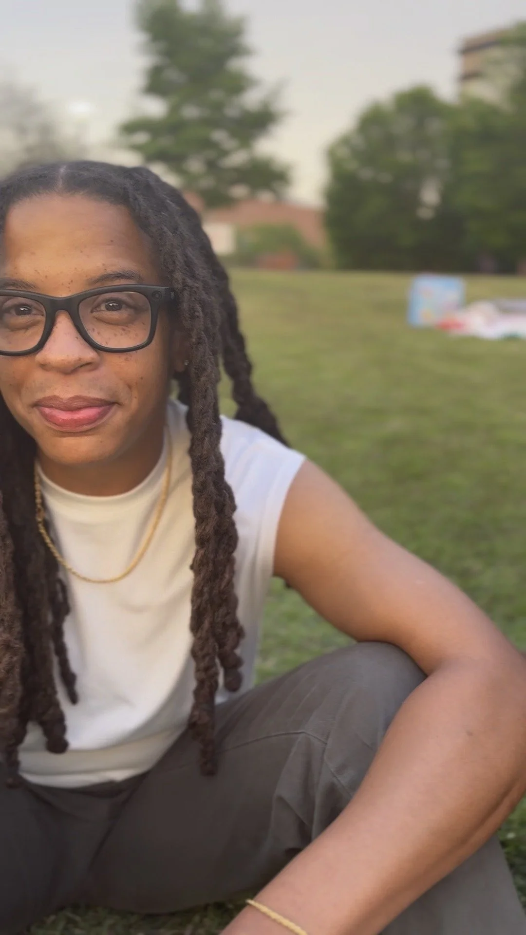 A young woman with braids, wearing glasses and a white sleeveless top, sitting outdoors on the grass with a blurred background of trees and buildings.