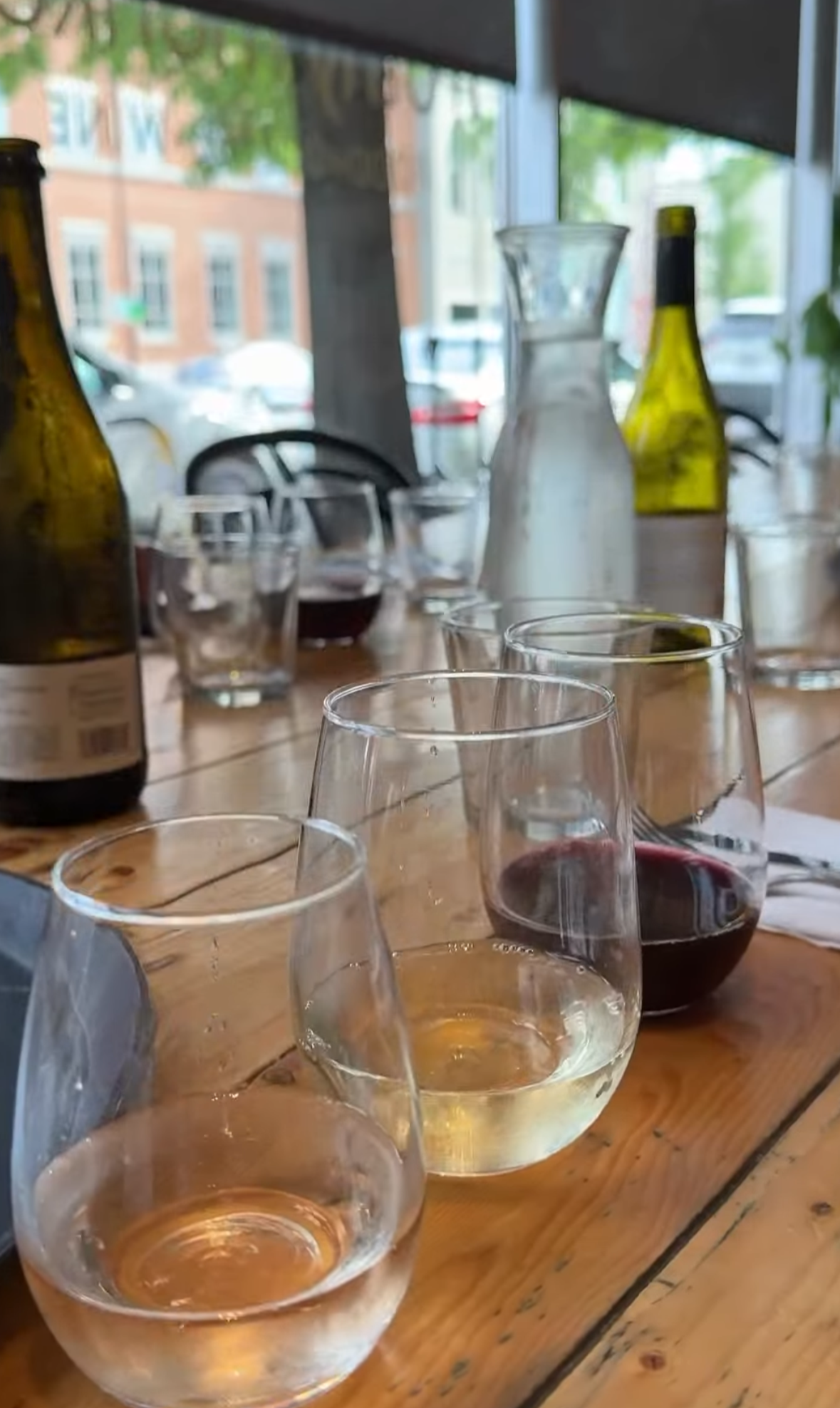 Multiple glasses of white wine and bottles on a wooden table inside a restaurant, with a large window showing cars and buildings outside.