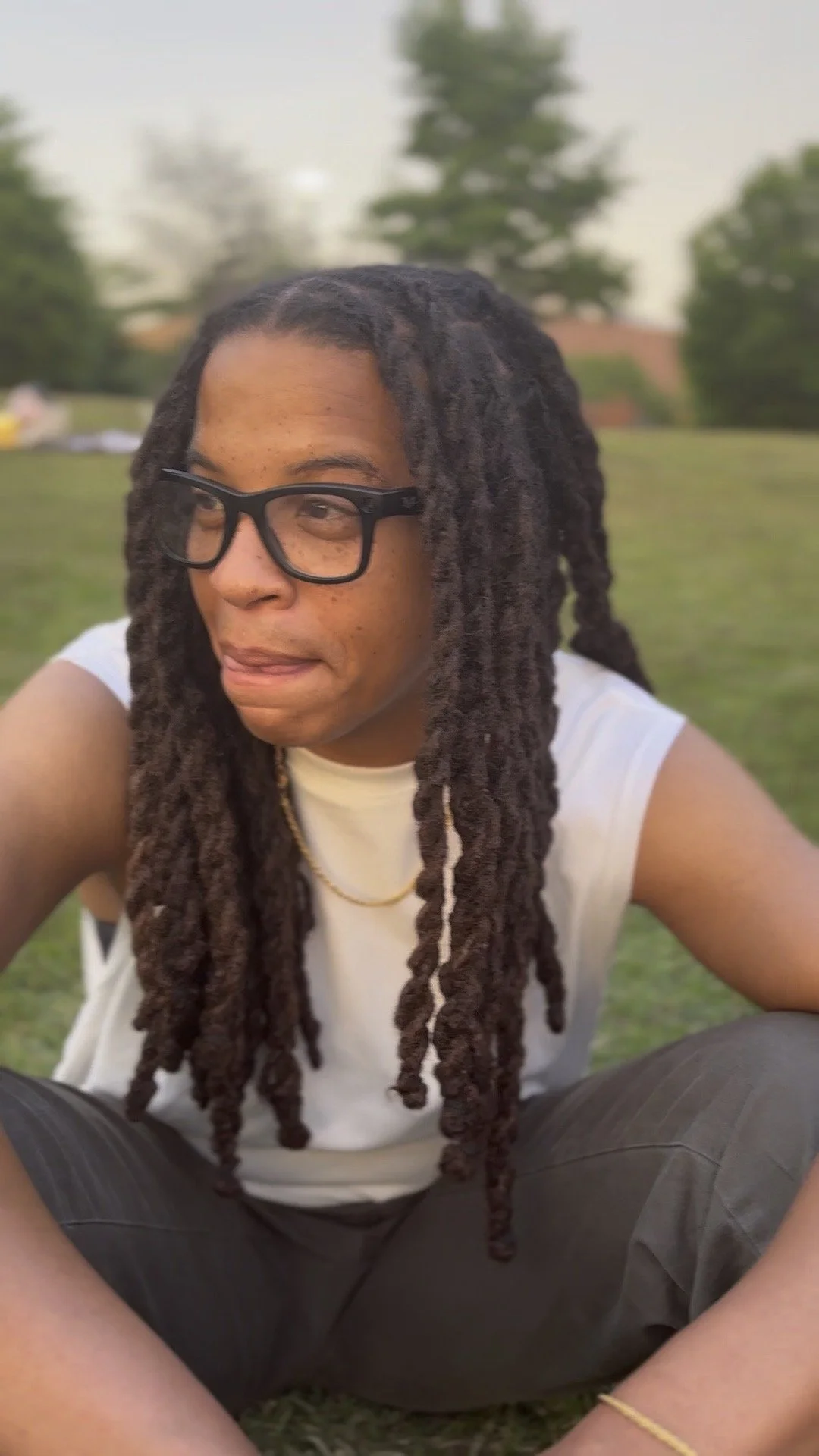 A person with long dreadlocks, wearing black glasses, a white sleeveless shirt, and a gold chain, sitting on the grass outdoors with trees in the background.
