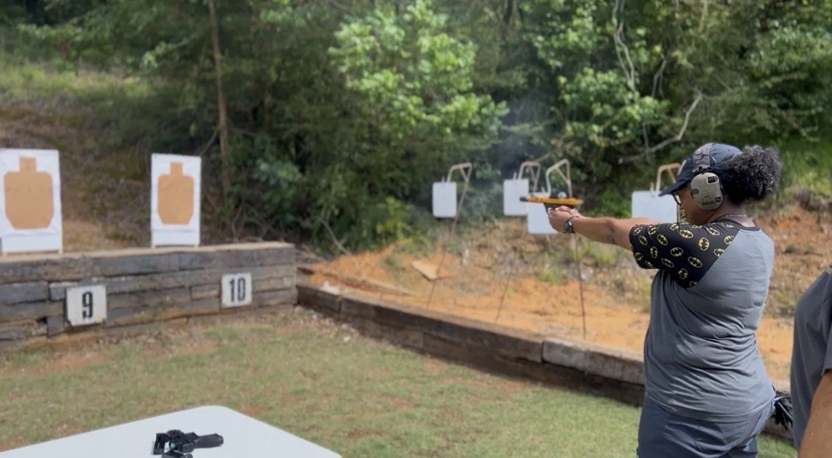 A woman shooting a handgun at outdoor shooting targets at a gun range. She is wearing safety headphones, a baseball cap, and a t-shirt with a Batman logo pattern, aiming the gun downrange.