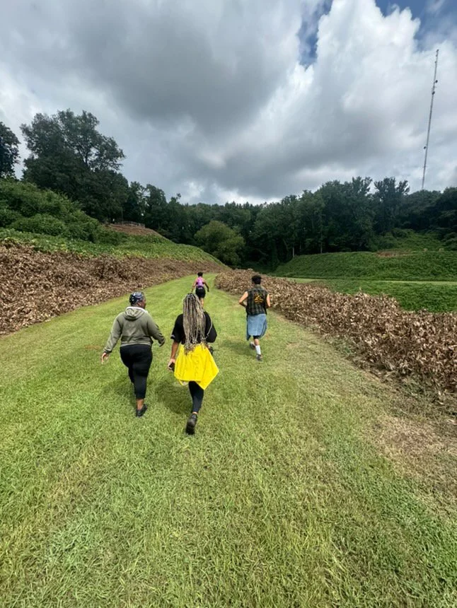 Four people are running along a grassy trail in a rural area with terraced fields on either side and trees in the background under cloudy skies.