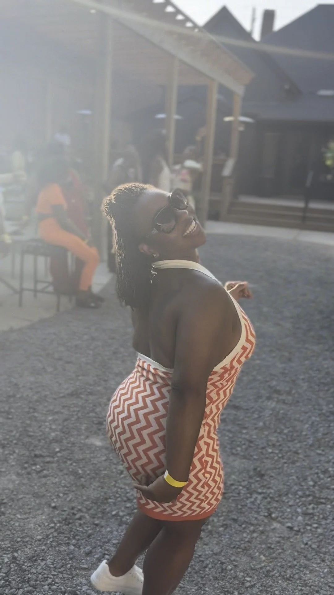 A woman smiling and posing outdoors in a zigzag patterned red and beige dress, wearing sunglasses, with people sitting and standing in the background, and sunlight creating a glow effect.