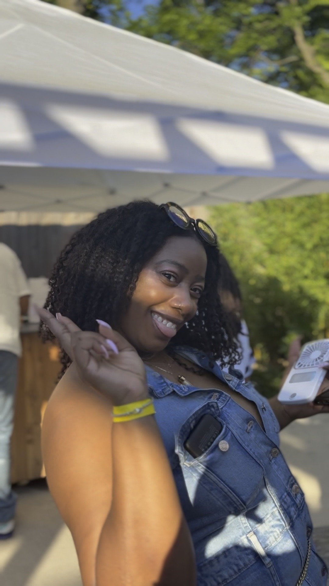A woman with curly hair smiling and making a peace sign at an outdoor gathering.