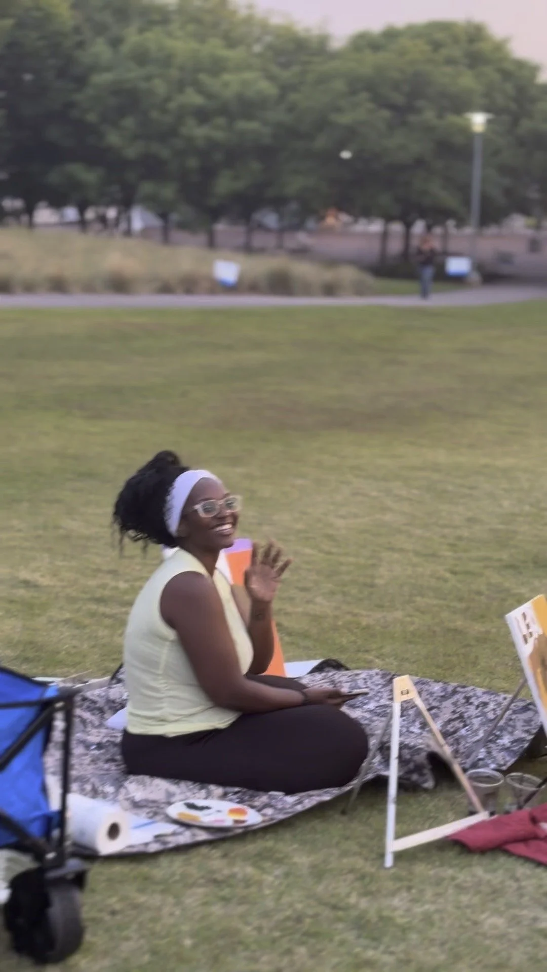 Smiling woman sitting on a blanket at an outdoor event, with art supplies and informational signs around her.