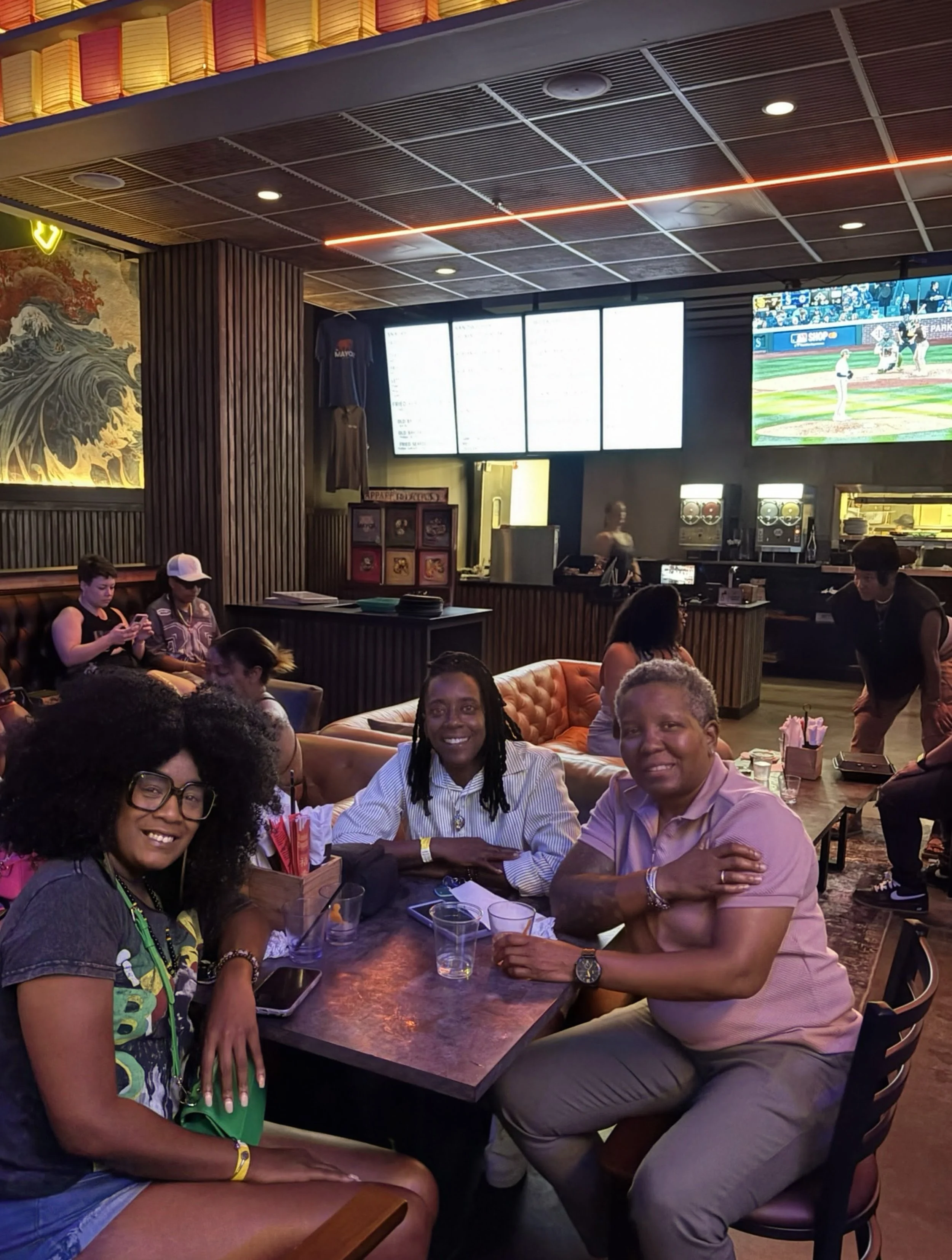 People sitting in a bar or restaurant, some are using phones, others are smiling and facing the camera, with a large screen displaying a baseball game in the background.