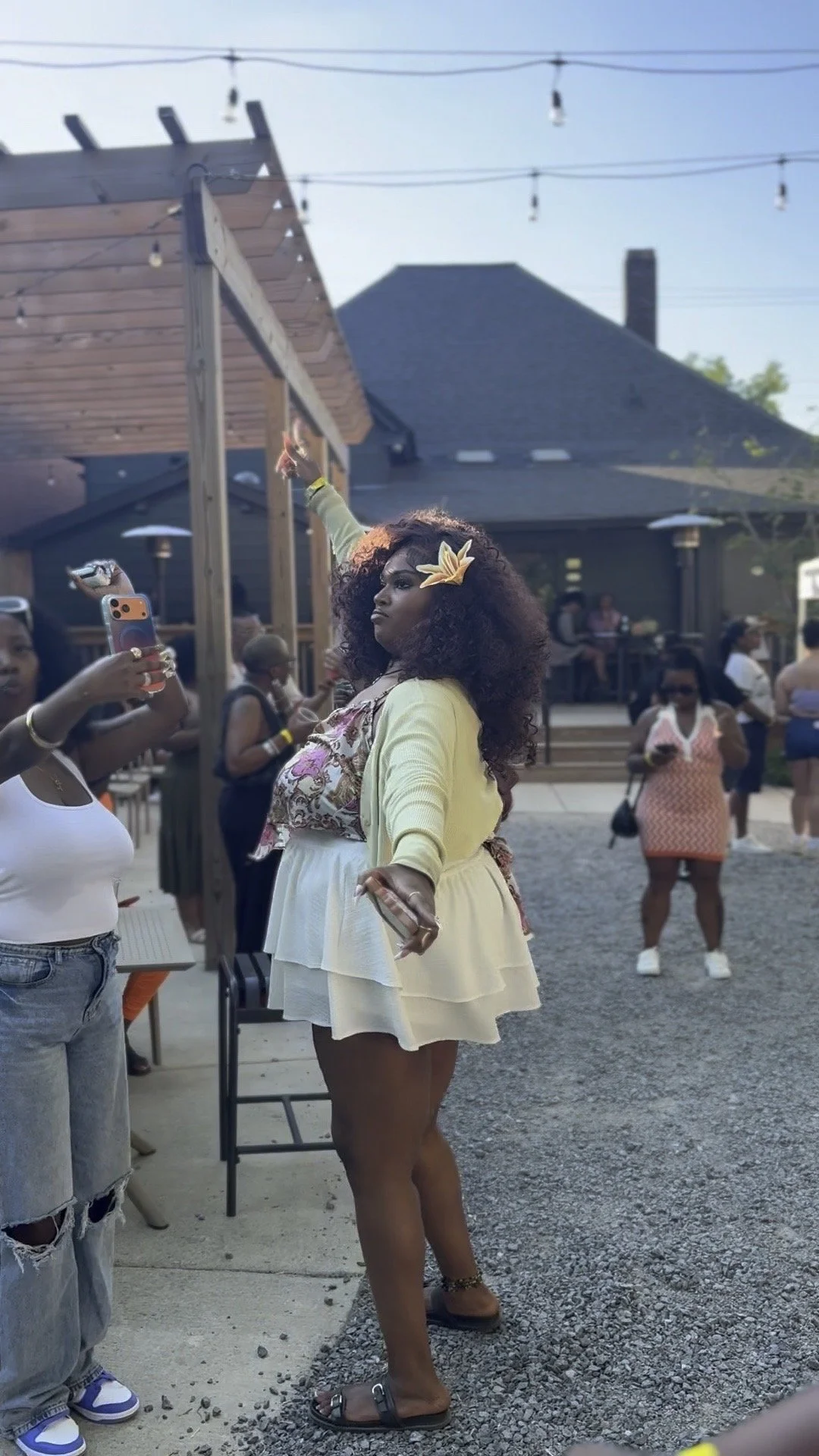 A woman with curly hair wearing a yellow cardigan and cream dress, standing outdoors, with people in background at a social gathering.