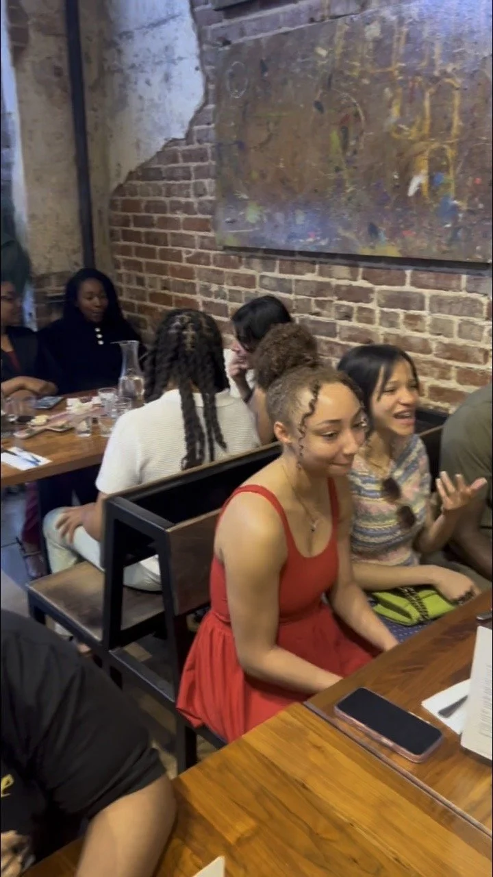 Group of diverse young women sitting at a wooden table in a restaurant with exposed brick walls, talking and smiling.