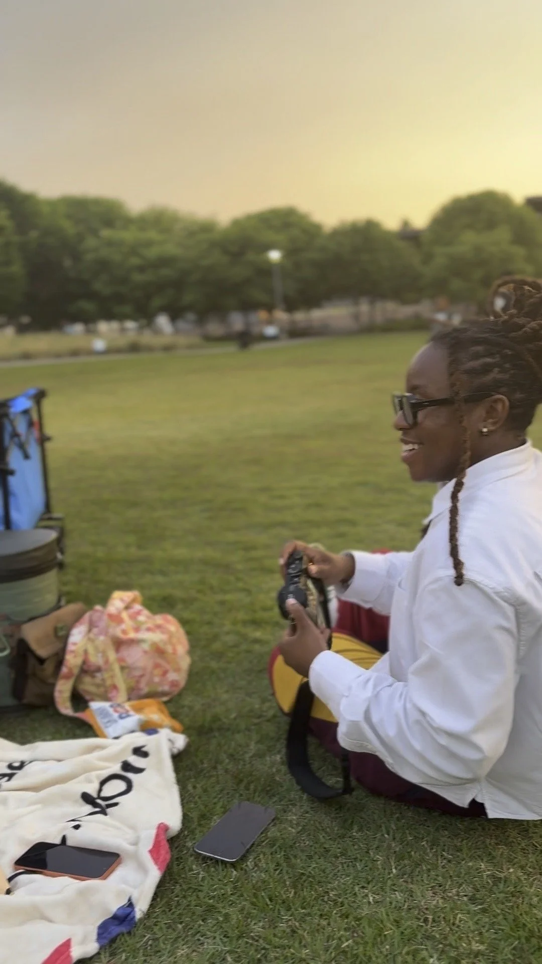 A woman with glasses and long hair styled in twists, sitting on grass with various bags and clothes, smiling and holding a camera, outdoor park setting with trees in the background.