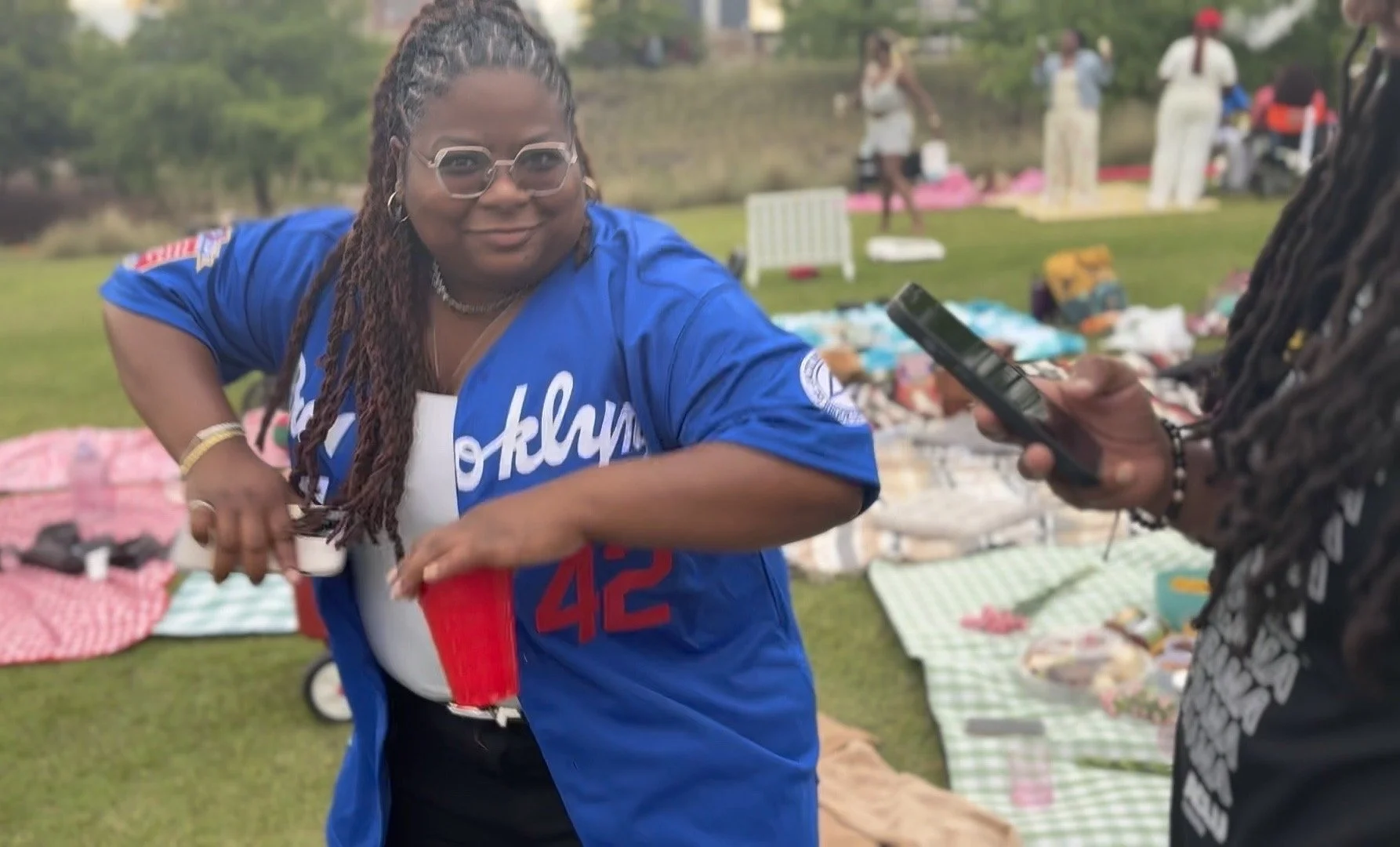 Woman in a blue sports jersey with glasses and long braids holding a red solo cup at outdoor gathering.