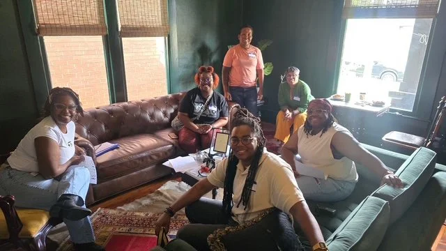 Group of six women in a living room, some seated on couches and one standing, with a woman seated at a desk, a laptop, and papers, during daytime with sunlight coming through windows.