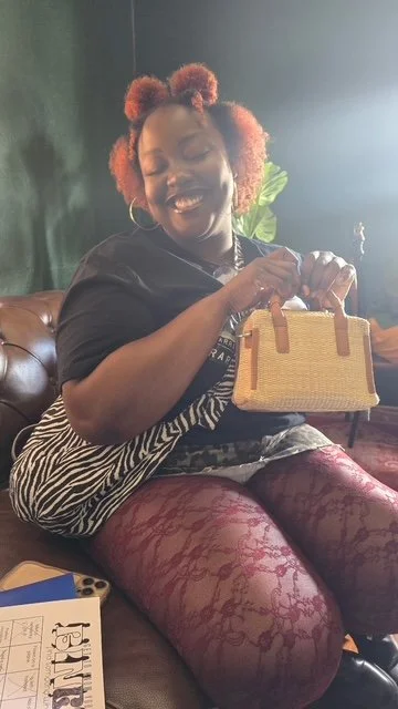 A woman with curly, reddish hair, sitting on a sofa, smiling and holding a small yellow handbag with brown handles, wearing a black top, zebra print skirt, and red patterned tights.