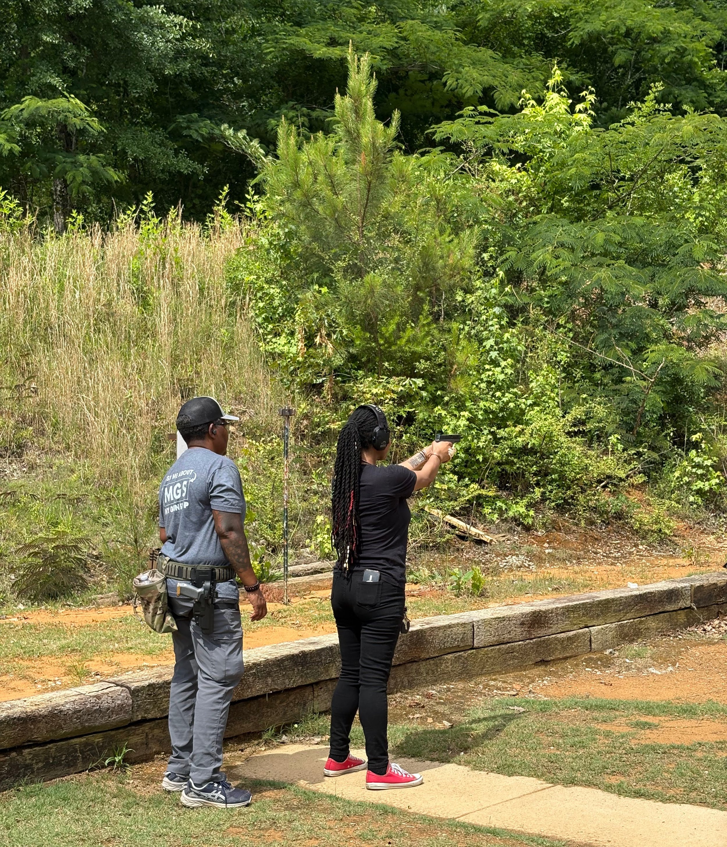 Three people standing outdoors near a wooded area, one person aiming a handgun while the other two observe, next to a small raised wooden fence and a sandy pathway.