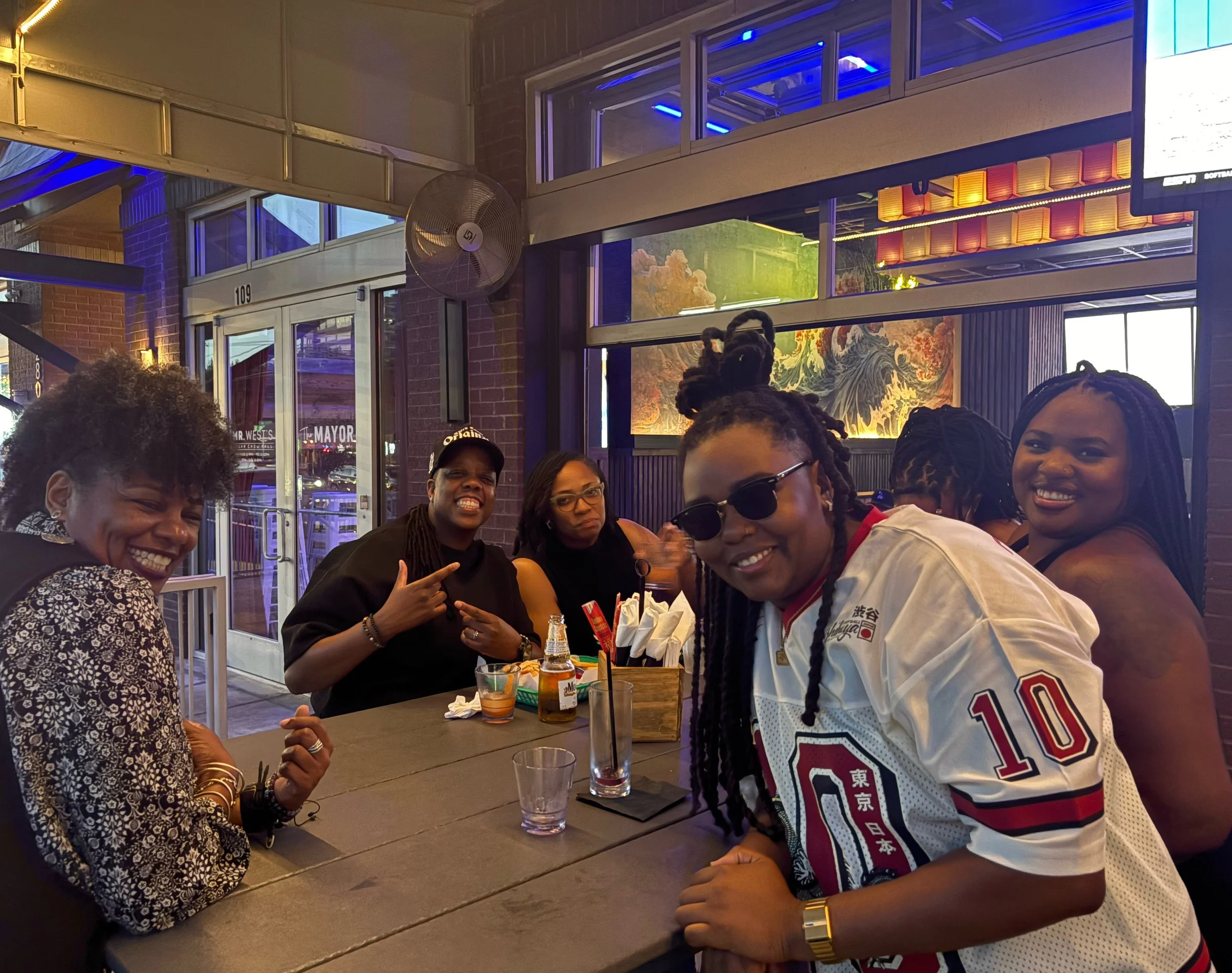 Group of people dining at a restaurant, smiling and posing for the photo, with drinks and utensils on the table, in a modern restaurant with decorative artwork and lighting.
