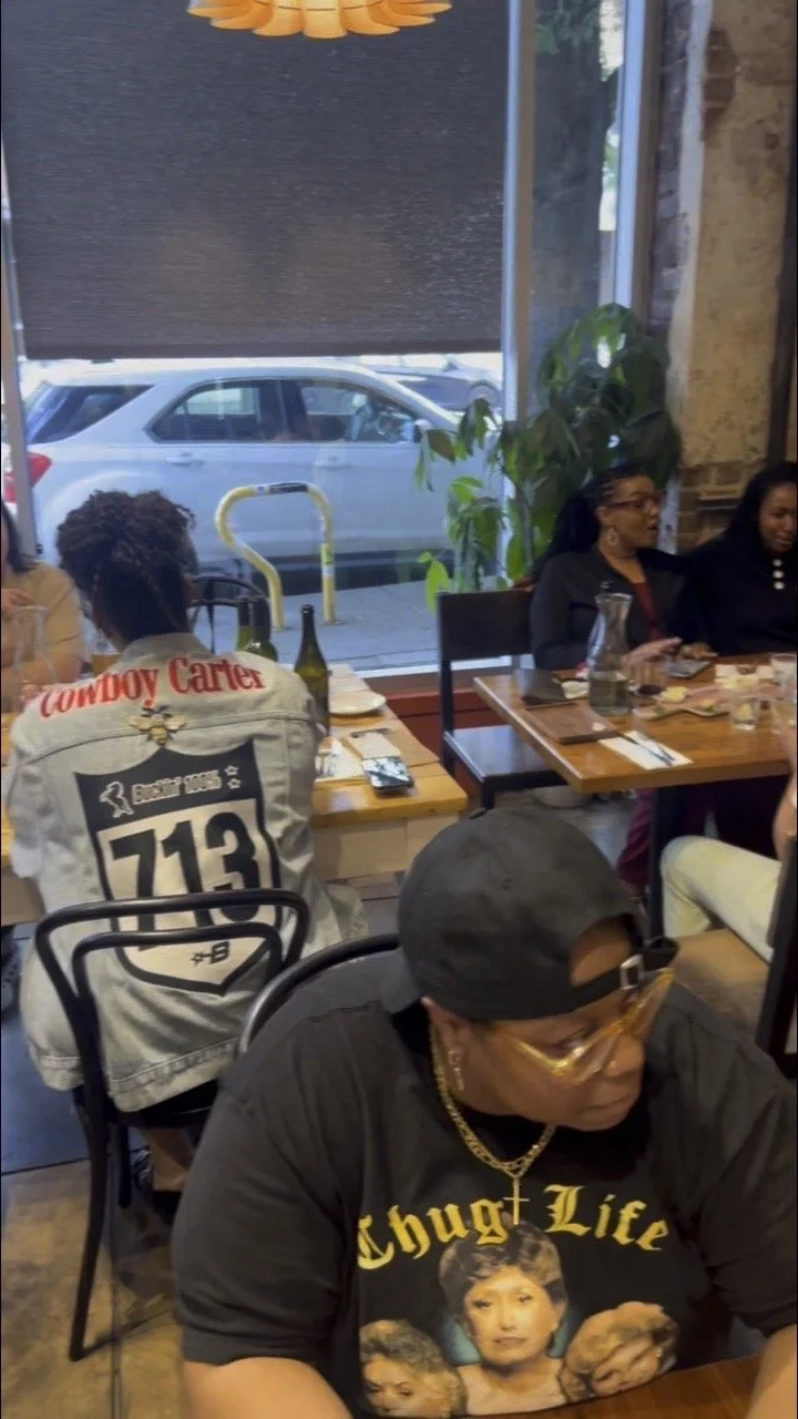 People dining inside a restaurant, with large front window showing parked cars outside. A woman in the foreground wears a black T-shirt with vintage women graphics and sunglasses, others are seated at tables, and a large green plant is near the windo