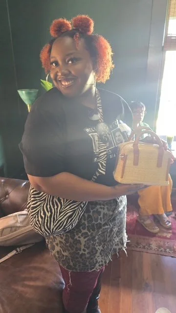 A woman with curly, reddish hair holding a tan handbag inside a room with dark walls and a window. She is smiling and wearing a black shirt with a graphic design and a zebra and leopard print skirt.