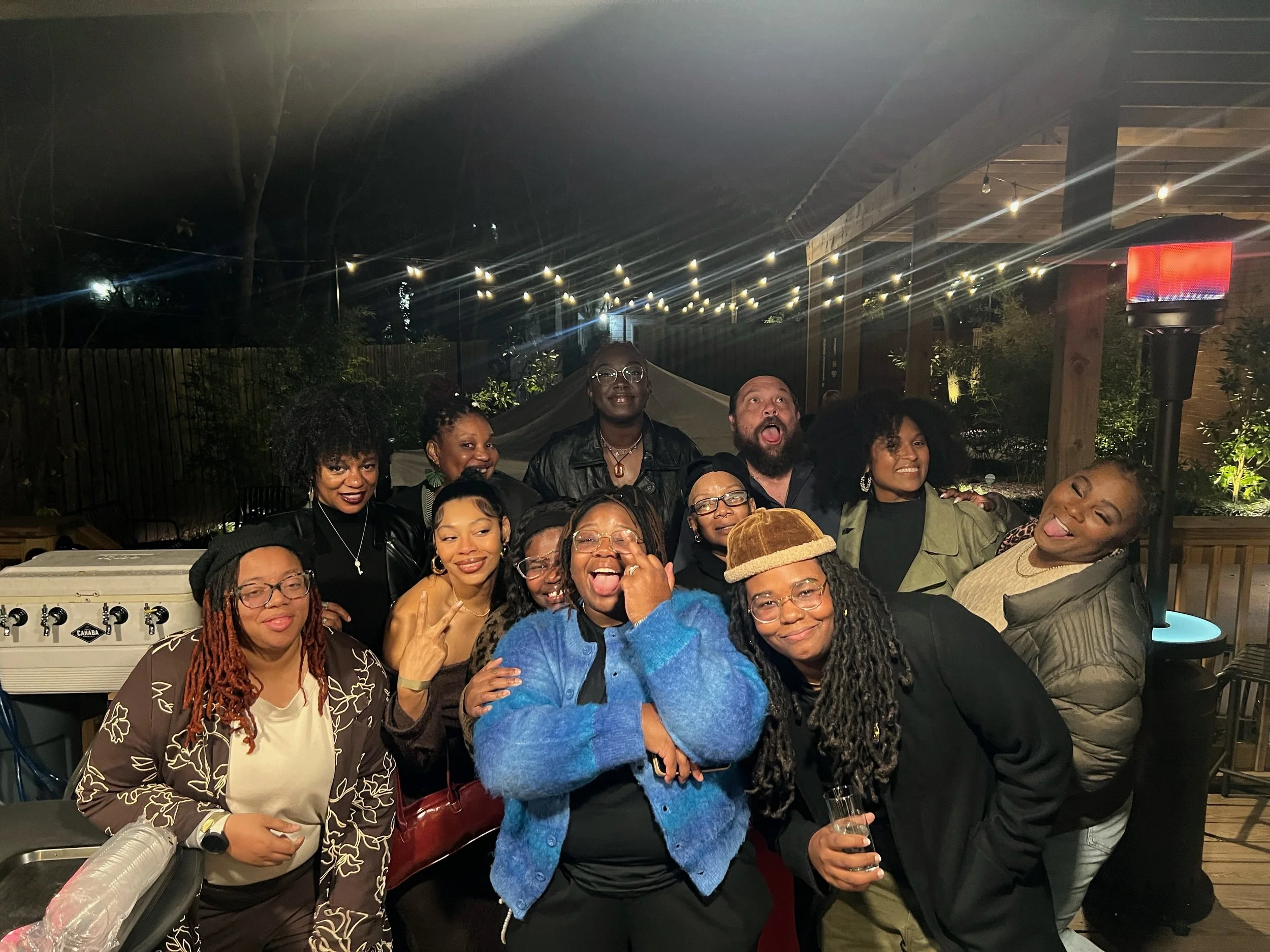 Group of people gathered outdoors at night, smiling and enjoying themselves, with string lights overhead and a red light in the background.