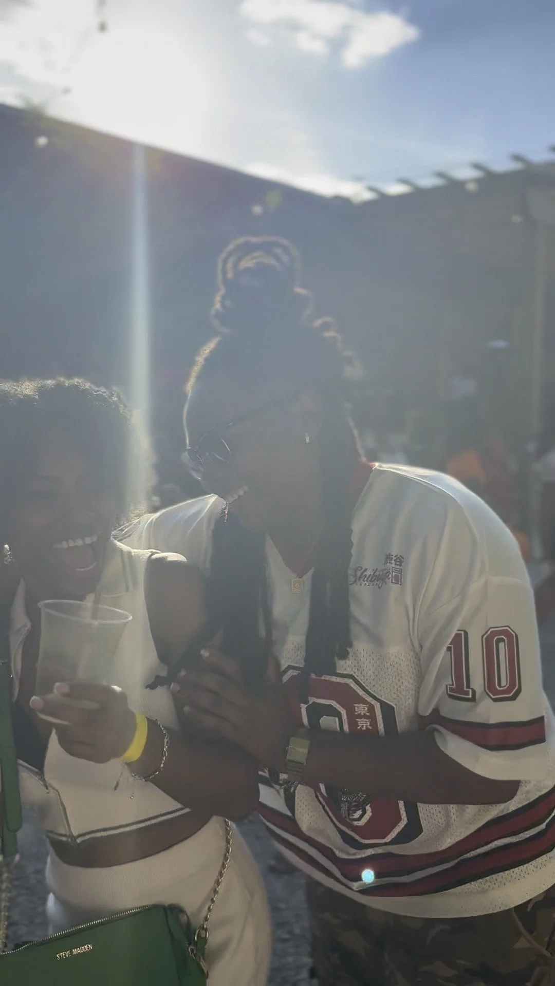 Two people standing side by side, one holding a drink with a straw, smiling and wearing a white sleeveless top, the other wearing a sports jersey with the number 10, sunglasses, and long black hair. Bright sunlight creates a backlit effect, causing t