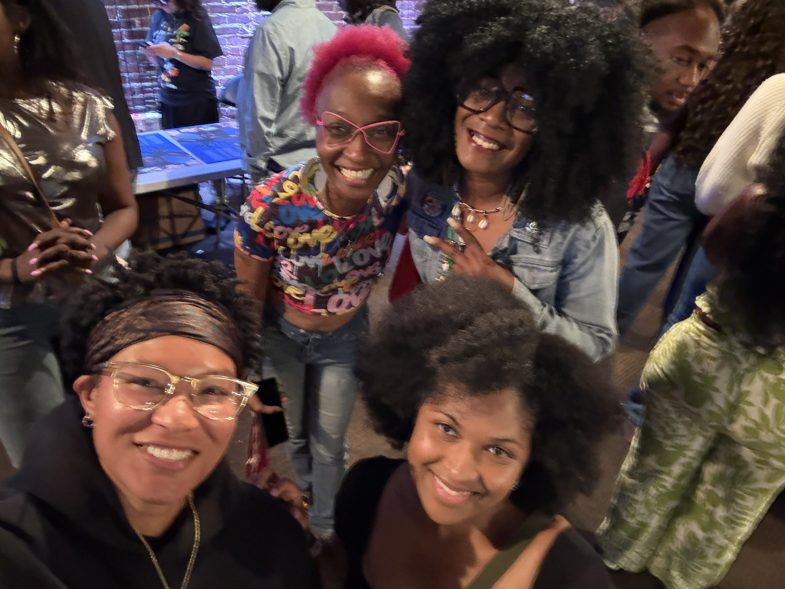 A group of smiling women taking a selfie in a dimly lit indoor setting. Some women have colorful hair and bold fashion styles, and there is a brick wall in the background.