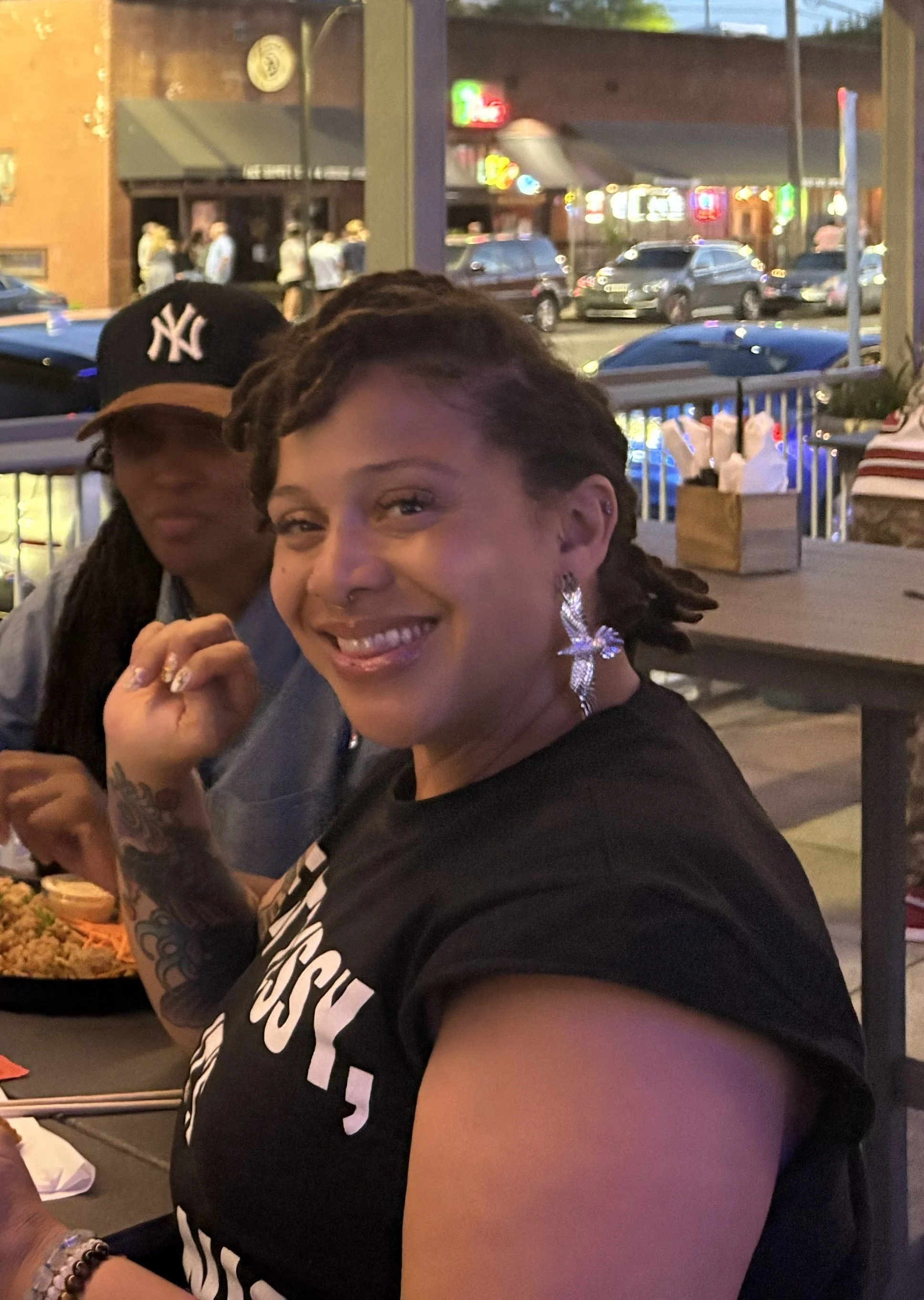 Two women at an outdoor restaurant at dusk, smiling and sitting at a table with a plate of food, with a background of parked cars and neon signs.