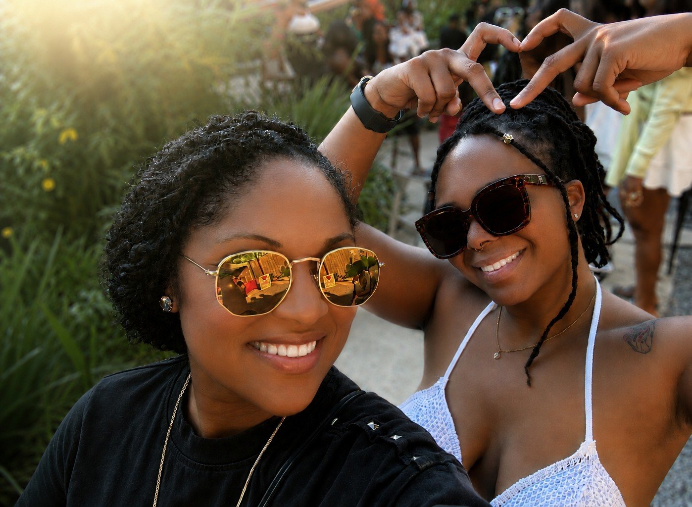 Two women taking a selfie at sunset, smiling, with gestures forming a heart shape above their heads. They are wearing sunglasses, with one in a black top and the other in a white top, surrounded by people in a park or outdoor setting.
