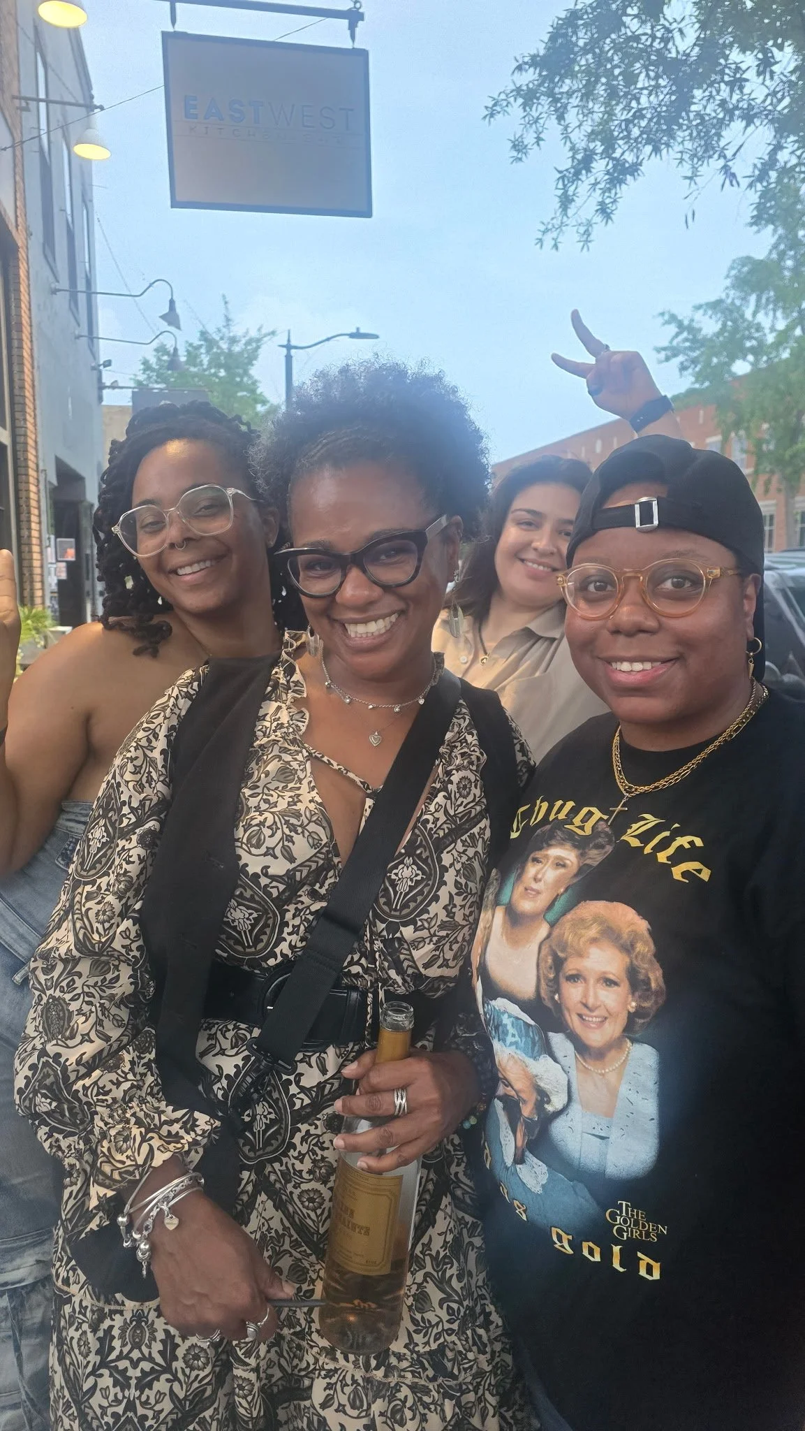 Four women smiling outdoors, one holding a bottle of wine, with trees and a street sign that says 'EASTWEST KITCHEN' in the background.
