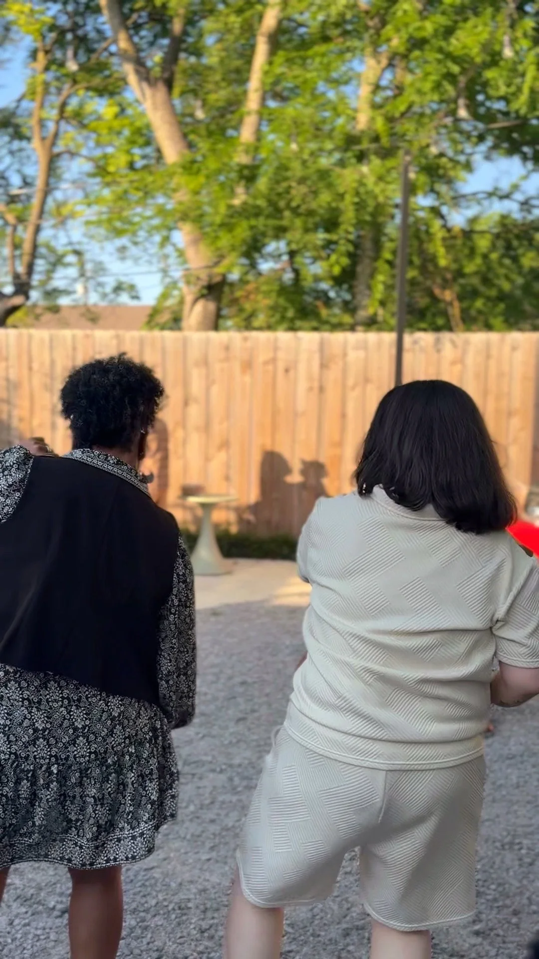 Two women sit outside on a gravel area, facing a wooden fence with trees behind it. One woman has short dark hair and wears a light-colored dress, while the other has curly dark hair and wears a patterned dress with a black vest. It appears to be a s