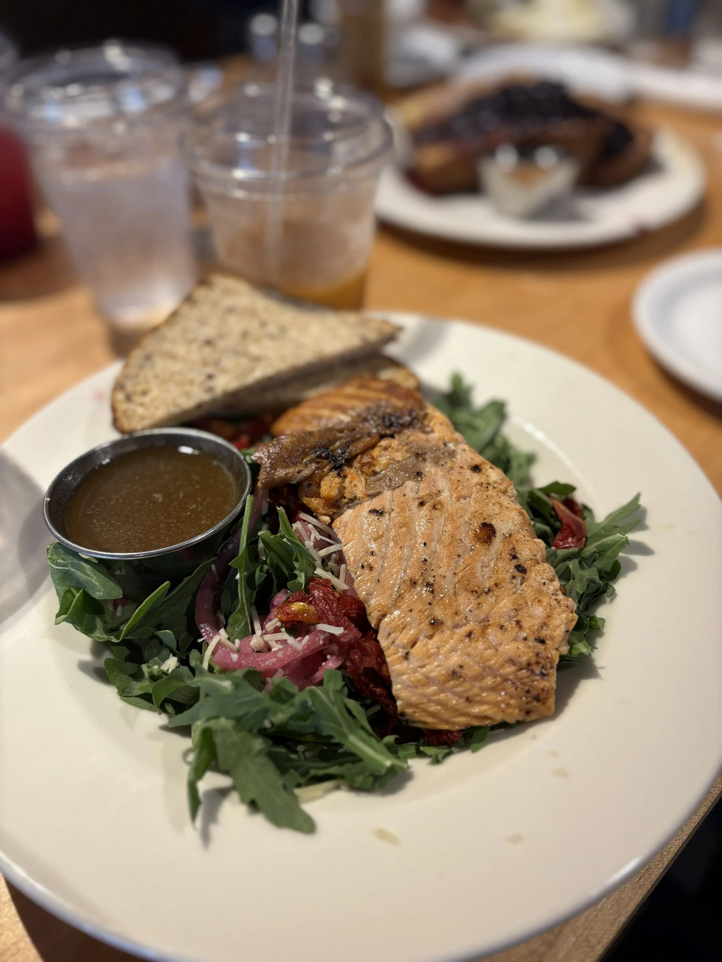 Grilled fish fillet served on a bed of mixed greens with a slice of multigrain bread, a small cup of sauce, and additional plates and drinks in the background.