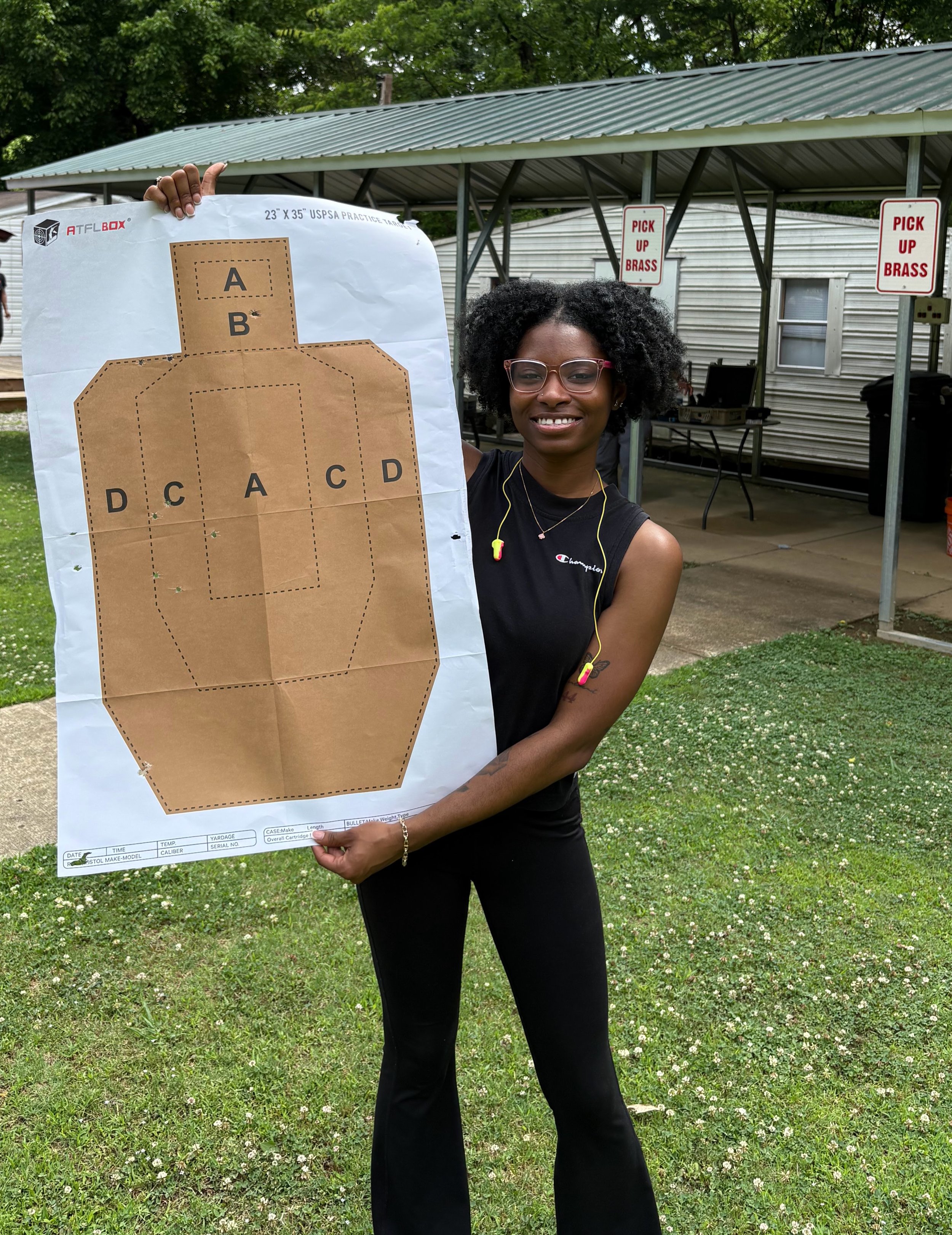 A woman with glasses smiling, holding a large shooting target poster with marked sections labeled A, B, C, and D, outdoors in a grassy area with trees and a shelter in the background, under cloudy weather.