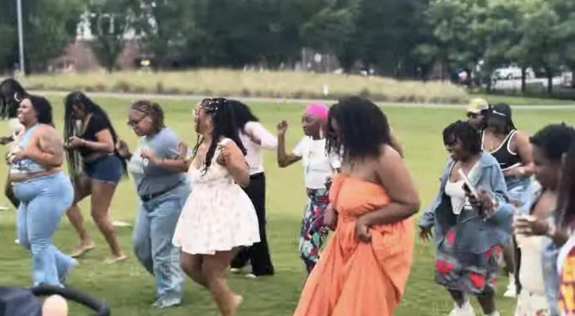 A group of diverse women dancing outdoors on a grassy field.