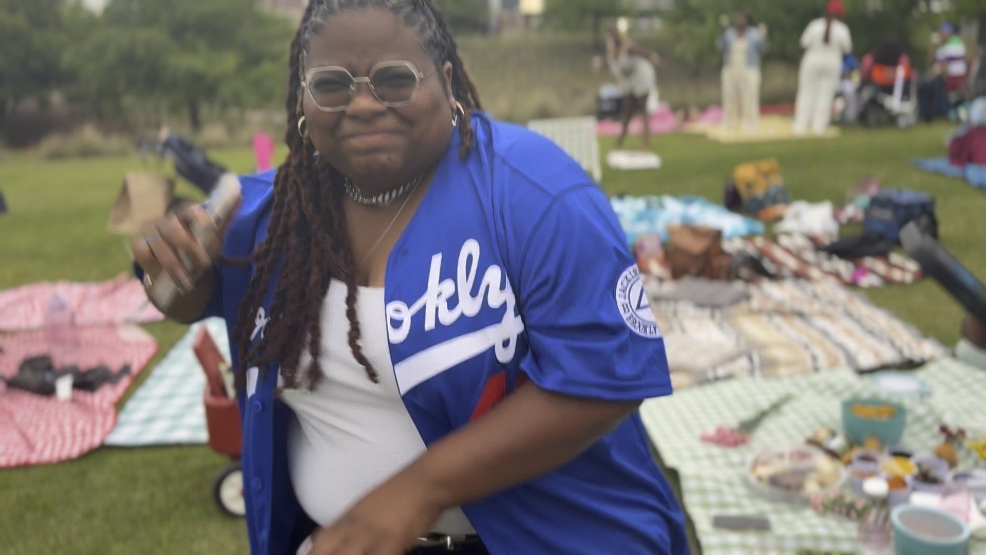 Woman in blue Oklahoma City Thunder jersey smiling and holding a drink at an outdoor yard sale with blankets and items on the grass in the background.