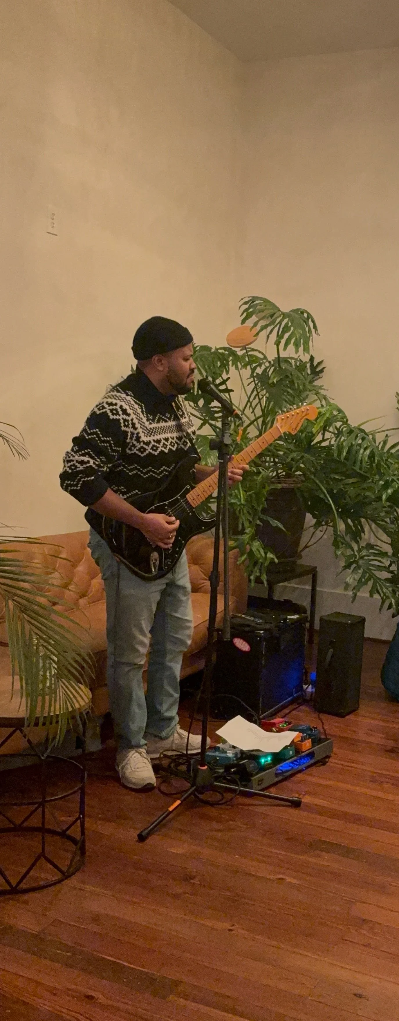 A man in a black beret, black and white patterned sweater, and light jeans playing an electric guitar and singing into a microphone in a cozy indoor setting with plants and warm lighting.
