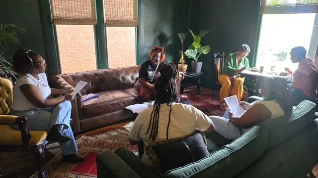 Group of people sitting and reading papers in a cozy living room with green walls and large windows.
