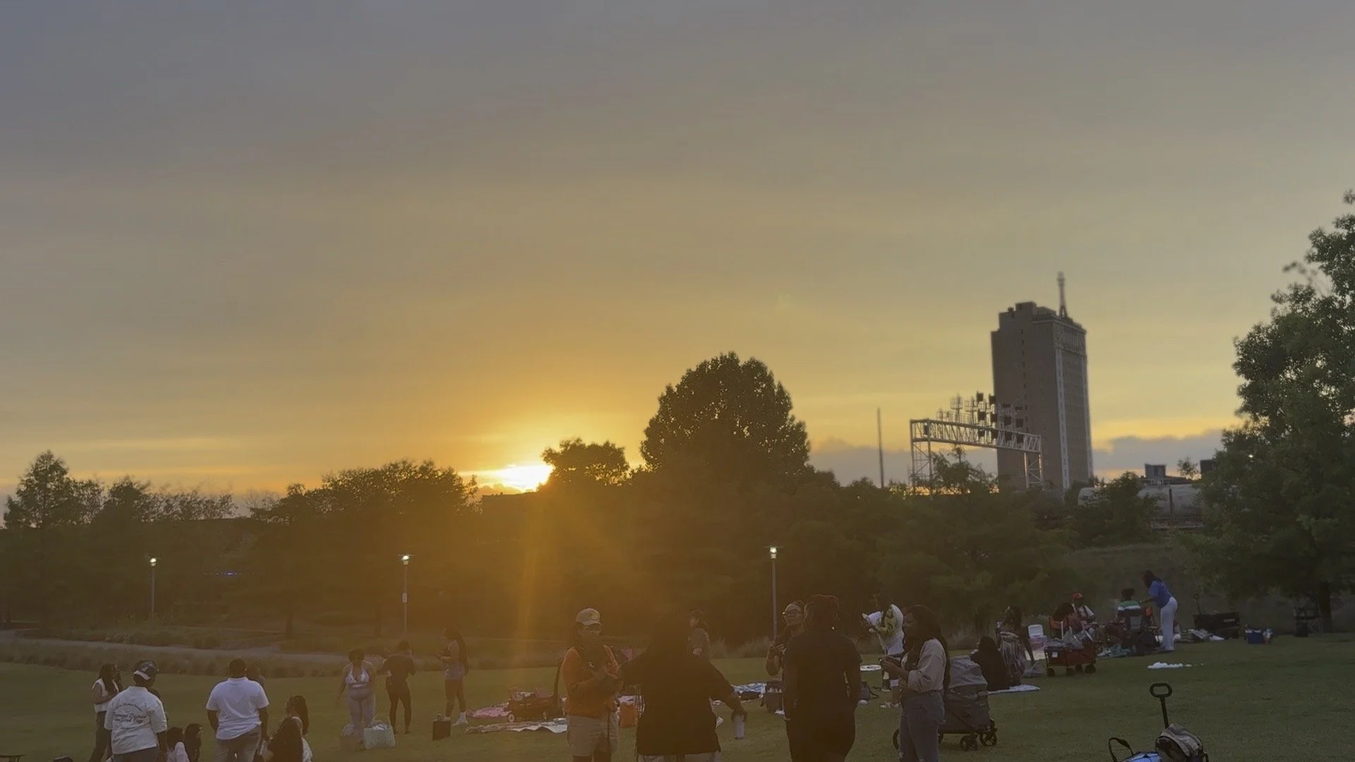 People gathered in a park during sunset with a cityscape and tall building in the background.