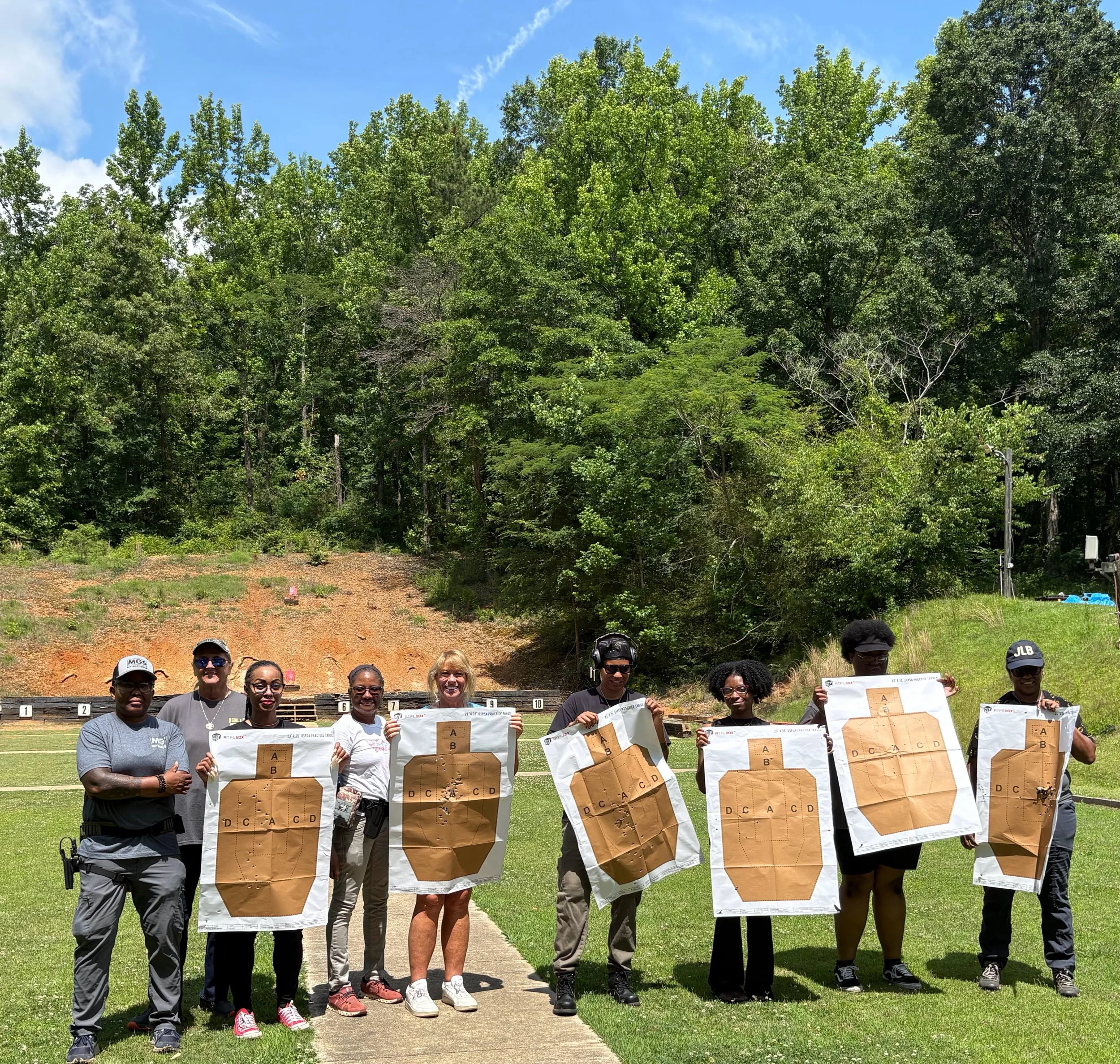 Group of people standing outdoors on a grassy field, holding large paper targets with lettered and numbered sections, with trees and a blue sky in the background.