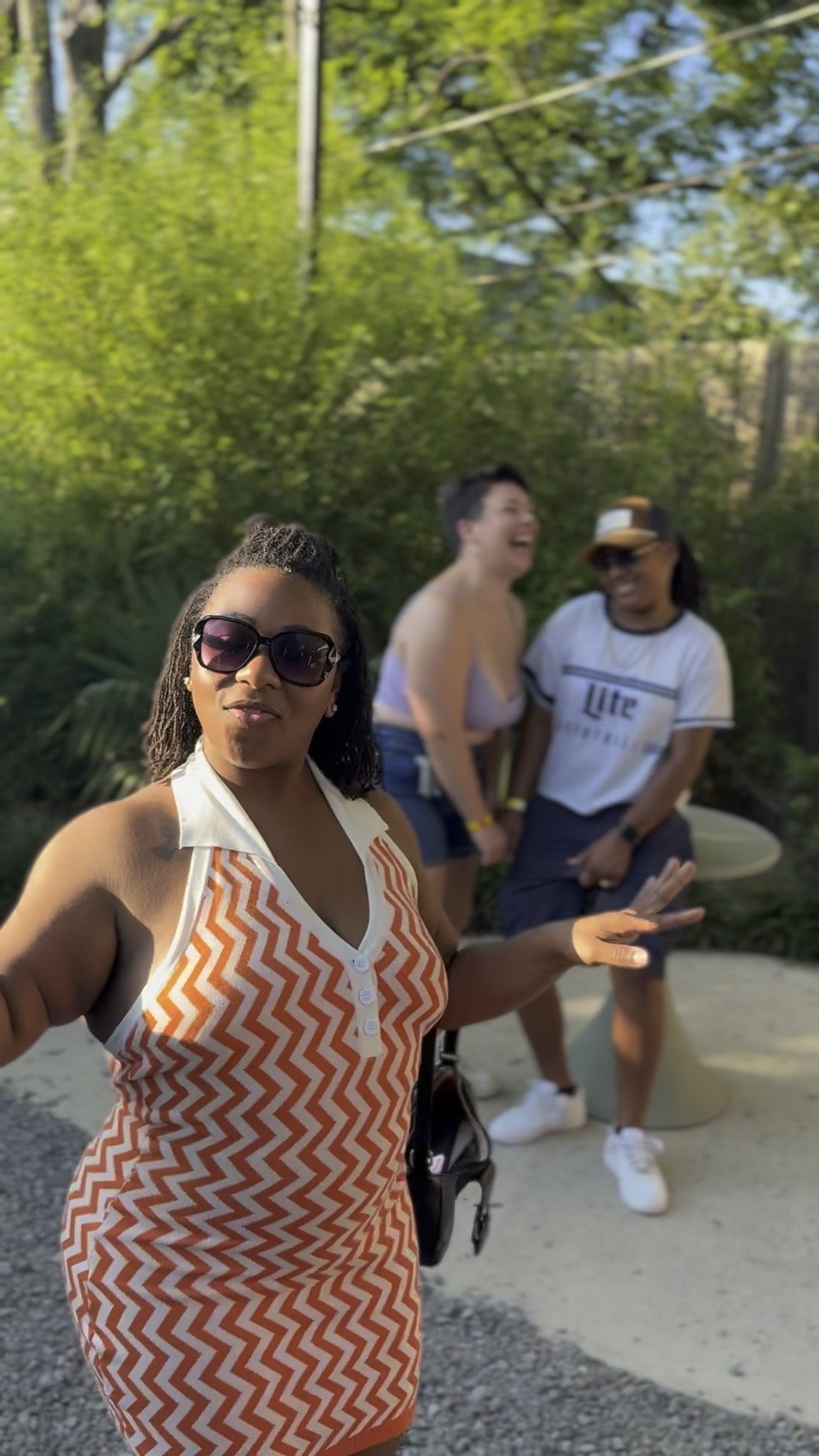 A woman wearing sunglasses and a sleeveless orange and white zigzag patterned dress is taking a selfie outdoors. Behind her, two women are walking and laughing, one with a flowy top and denim shorts, the other in a white shirt, black shorts, a tan bu
