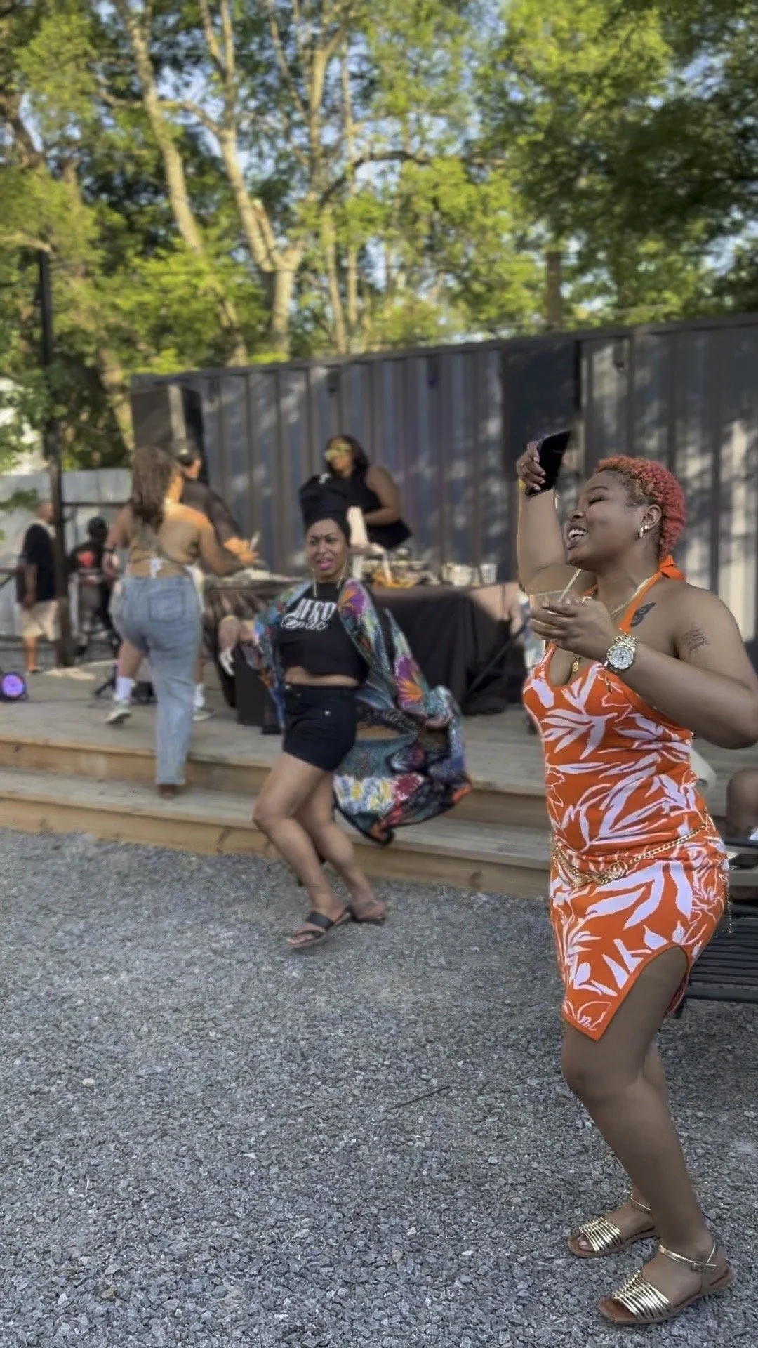 People enjoying an outdoor gathering, with a woman in a brightly patterned dress dancing and smiling as others socialize in the background near a food table.