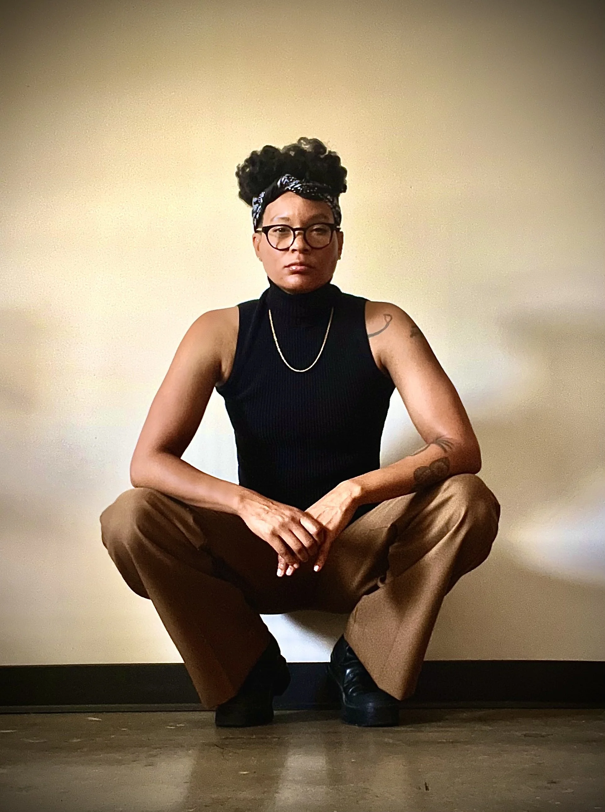 A person sitting on the floor with crossed arms, wearing glasses, a black sleeveless turtleneck shirt, beige pants, and black shoes, with a tattoo on the right arm and a headscarf. The background is a plain beige wall.