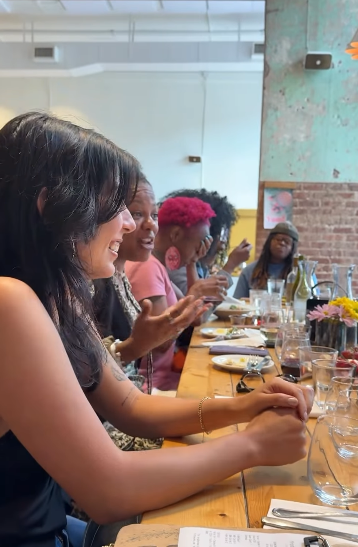 A group of women sitting at a long wooden table, engaged in conversation and using their phones, with plates, glasses, and flowers on the table inside a restaurant or cafe.