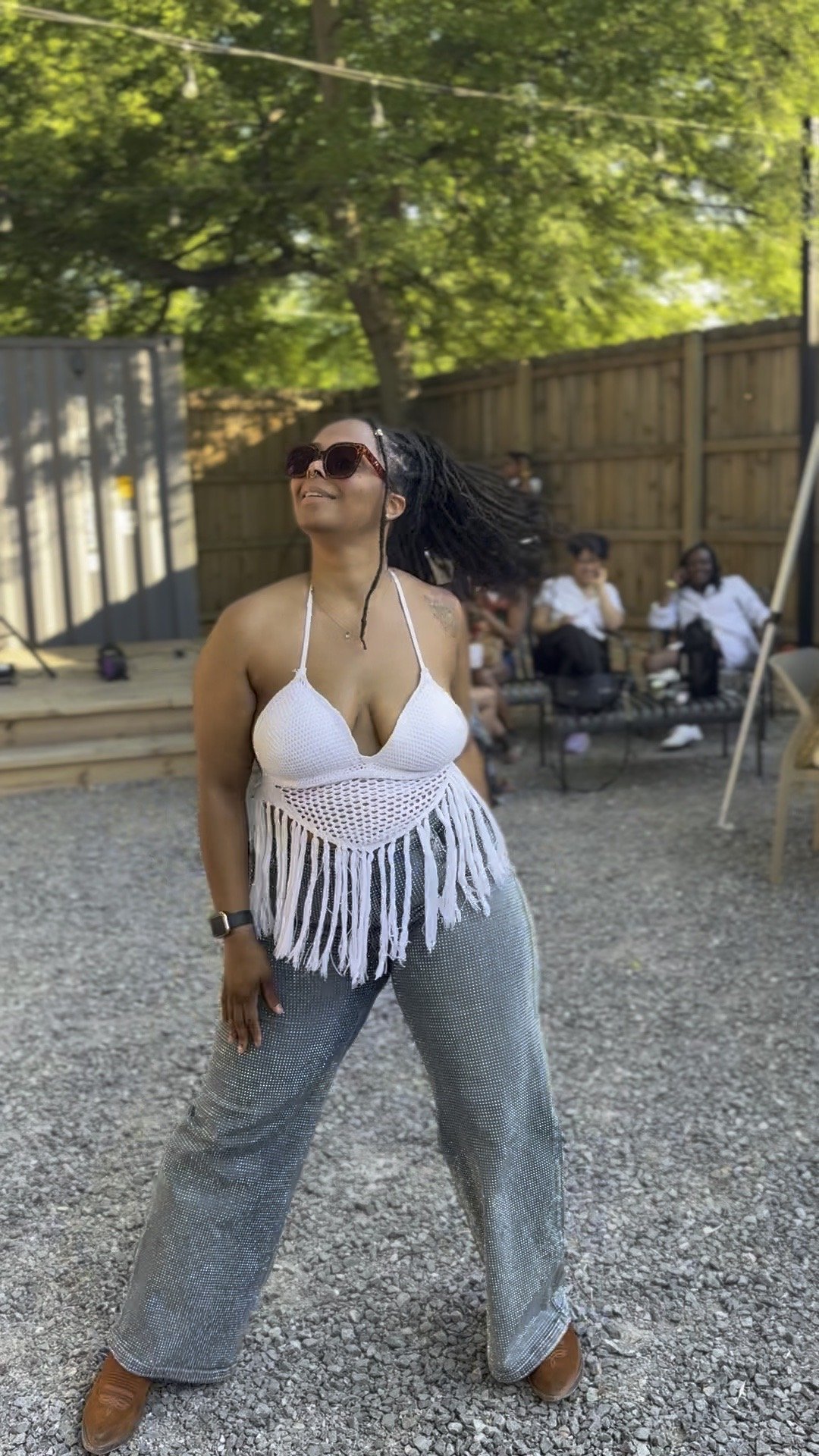 Woman in a white fringed top and patterned pants standing outdoors, smiling, with a group of people sitting on a bench in the background.