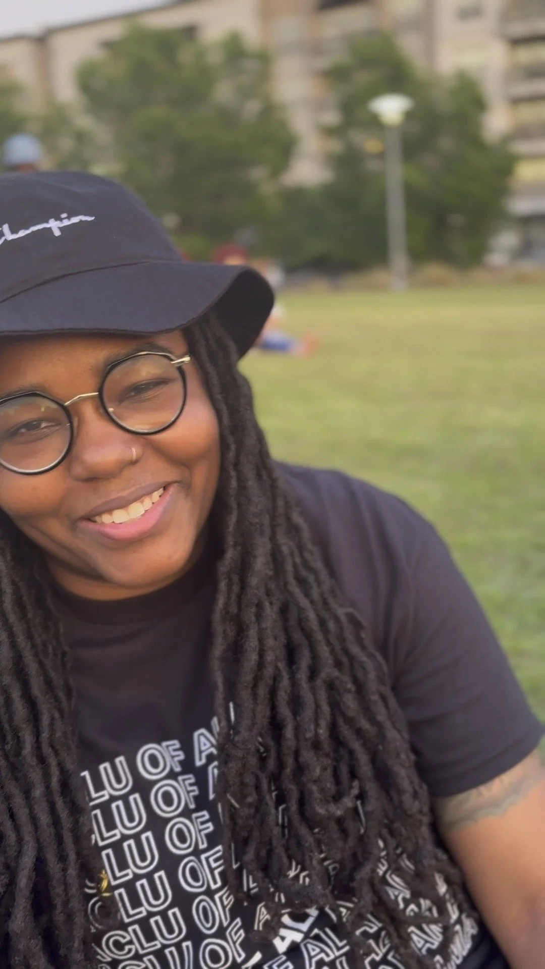 A smiling person with glasses, dreadlocks, a black cap, and a black shirt, outdoors in a park.