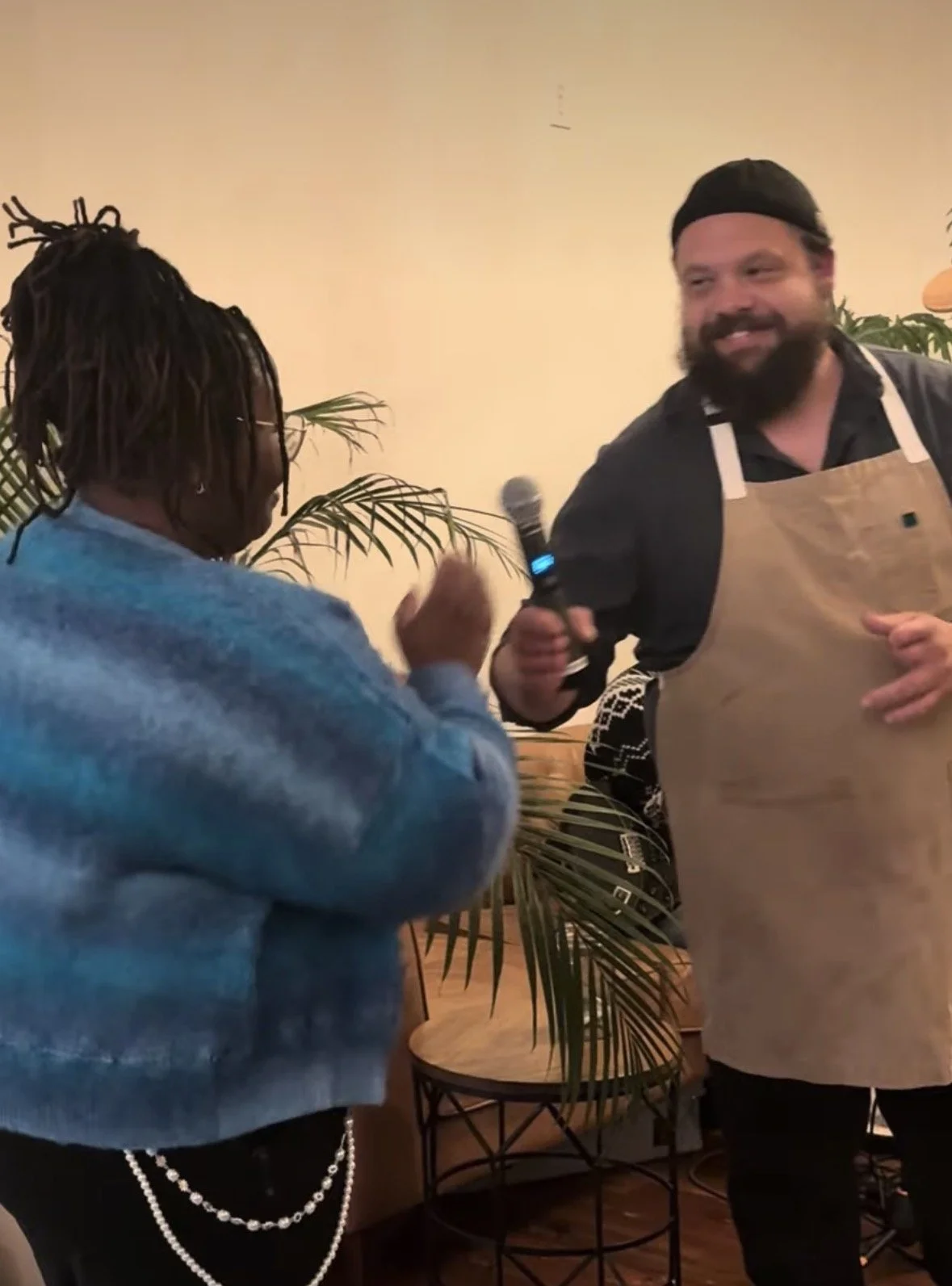 Two men are interacting indoors, one is holding a microphone and smiling, wearing an apron and a black cap, the other is talking with hand gestures, wearing a sweatshirt with pearl jewelry, with plants and a small table in the background.