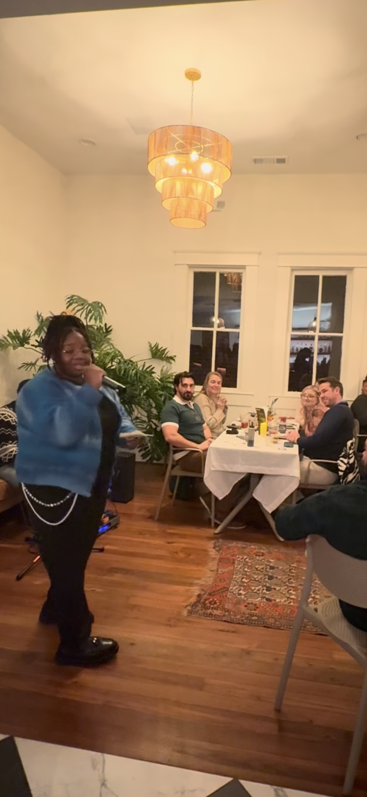 A woman with dreadlocks singing into a microphone at a dinner party, with guests seated around a table in the background.