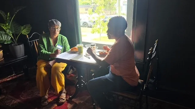 Two women sitting at a table in a cozy cafe with a large window, having drinks and conversation.