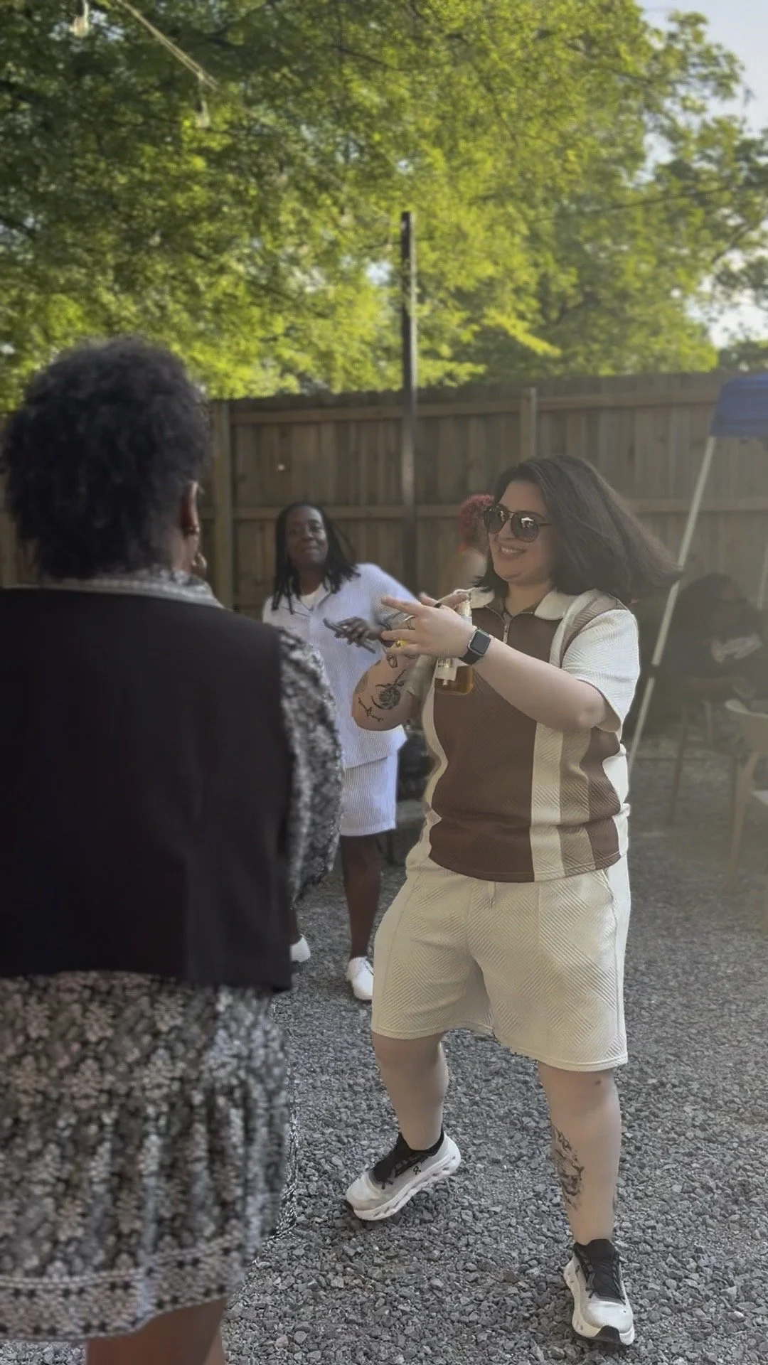 A woman with dark hair, sunglasses, and tattoos on her arm is smiling and pointing at another person during an outdoor gathering. Another woman with dreadlocks in the background is wearing a white striped outfit, and other people are present in the b
