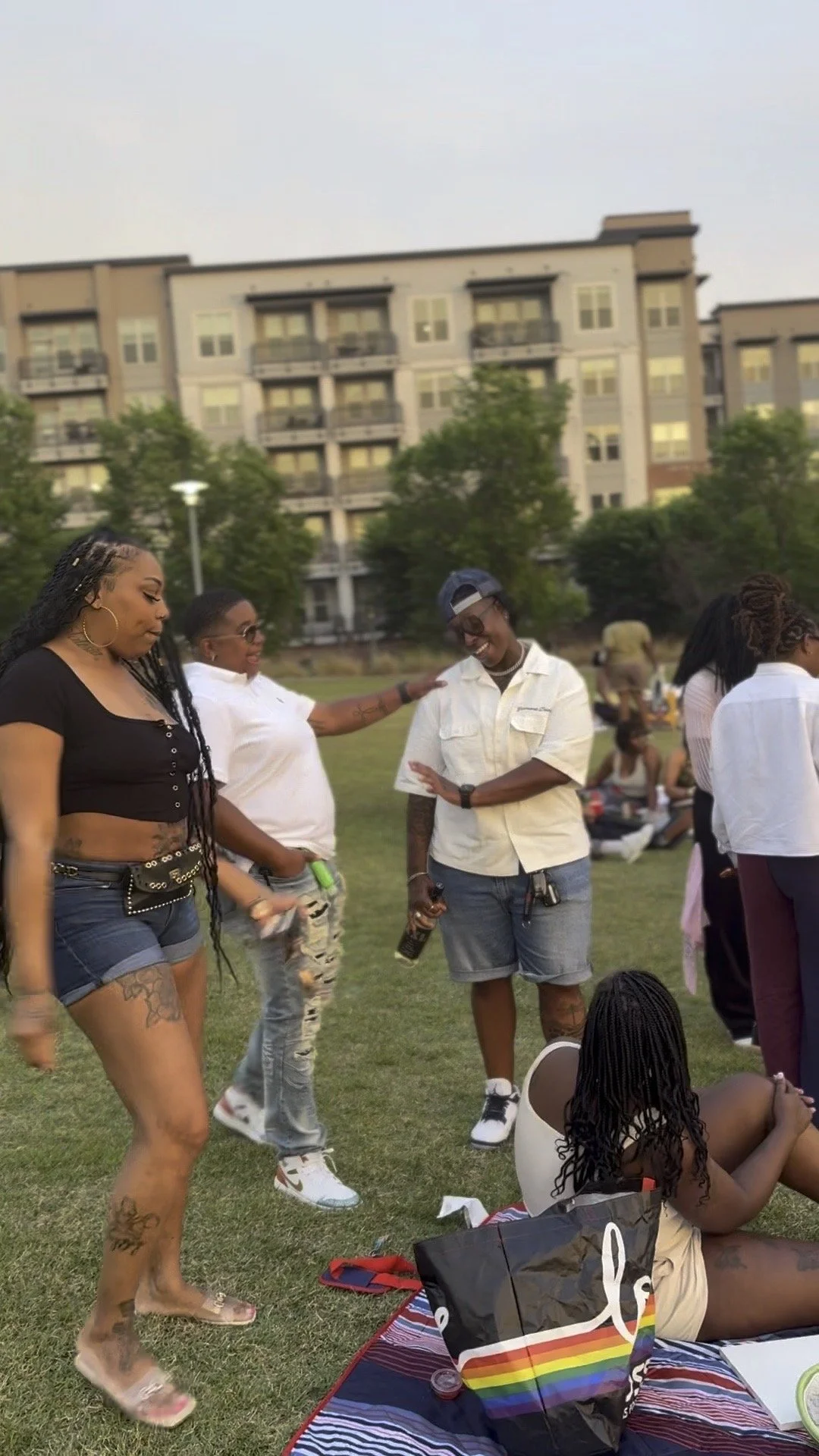 Group of people socializing outdoors on a grassy field with trees and an apartment building in the background, some sitting on a blanket, some standing and talking, in warm weather.