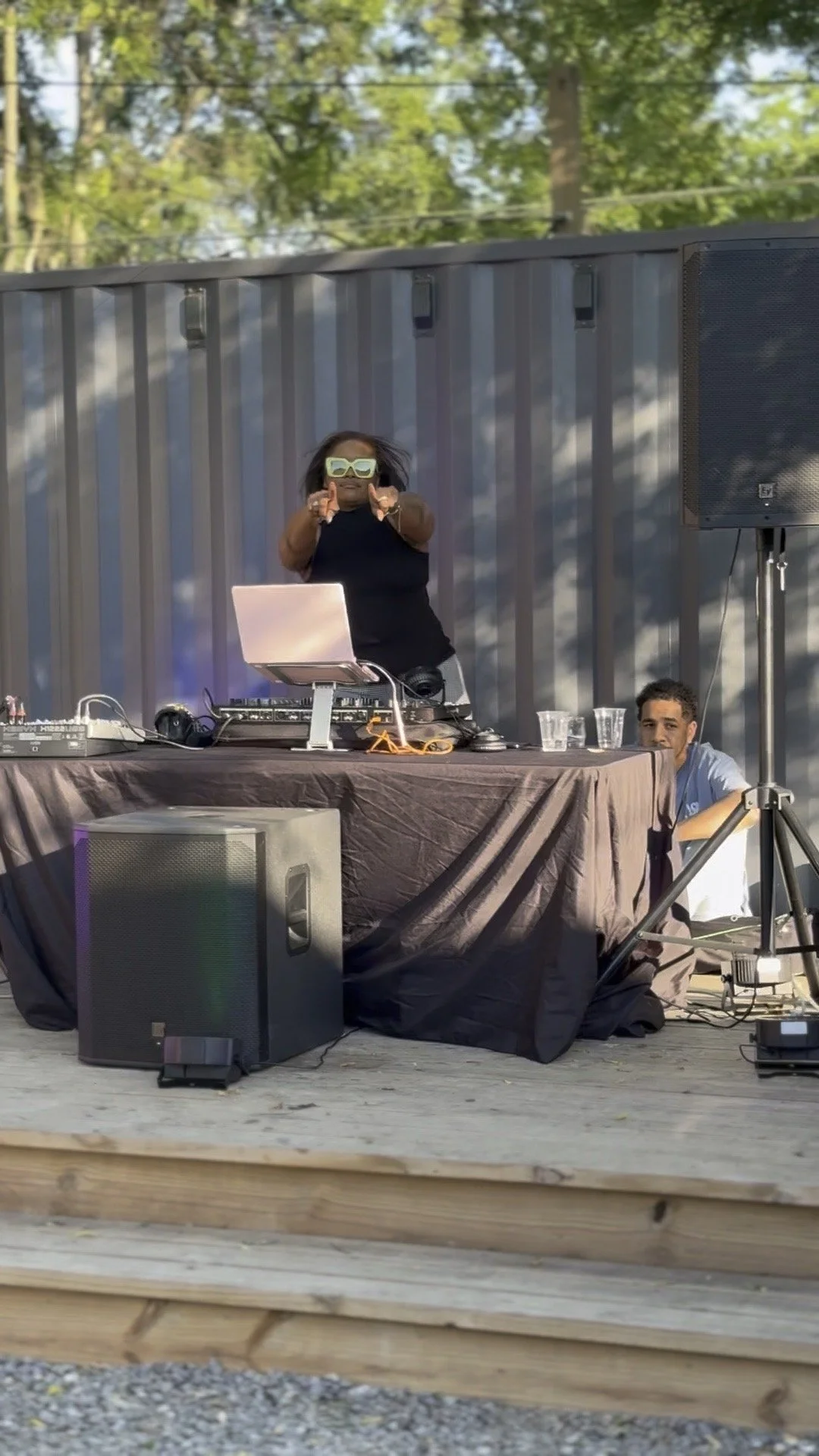 A DJ performing at an outdoor event, wearing large sunglasses and pointing at the camera, with audio equipment and a person sitting beside the table.